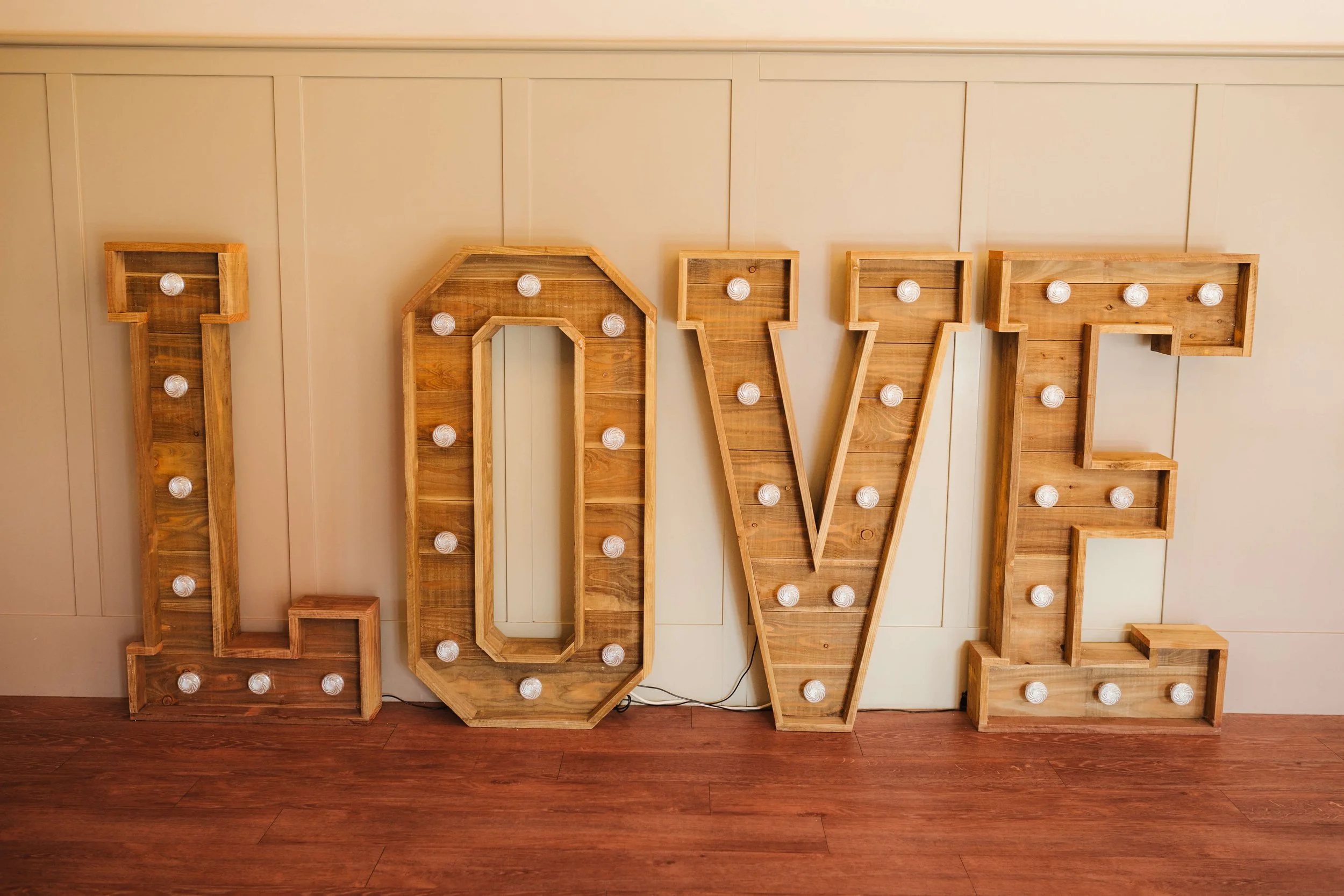 Large wooden marquee spelling out the word "LOVE" with white light bulbs inside each letter, placed against a beige wall with a wooden floor.