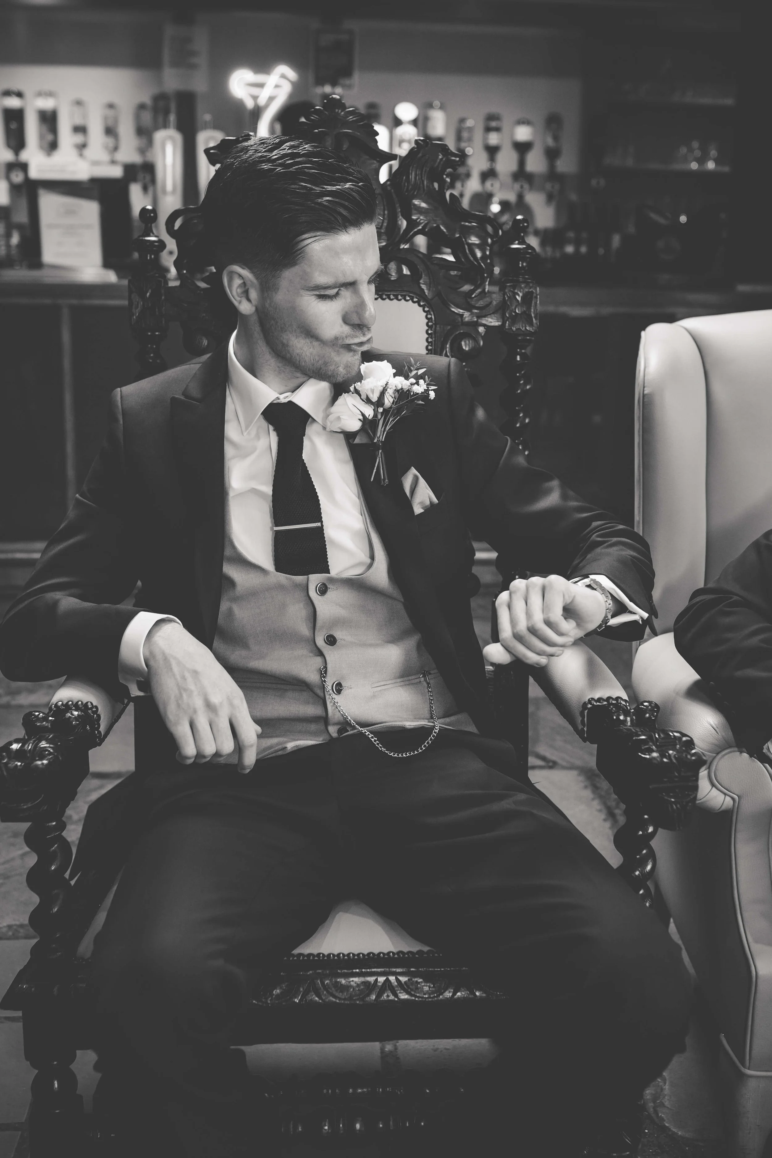 A groomsman dressed in a tuxedo with a boutonniere is sitting in a vintage chair and looking at his watch in a bar or lounge setting at wedding.