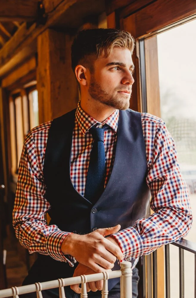 A young man with styled hair and a beard, dressed in a checkered shirt, vest, and tie, looking thoughtfully out a window inside a rustic wooden building.