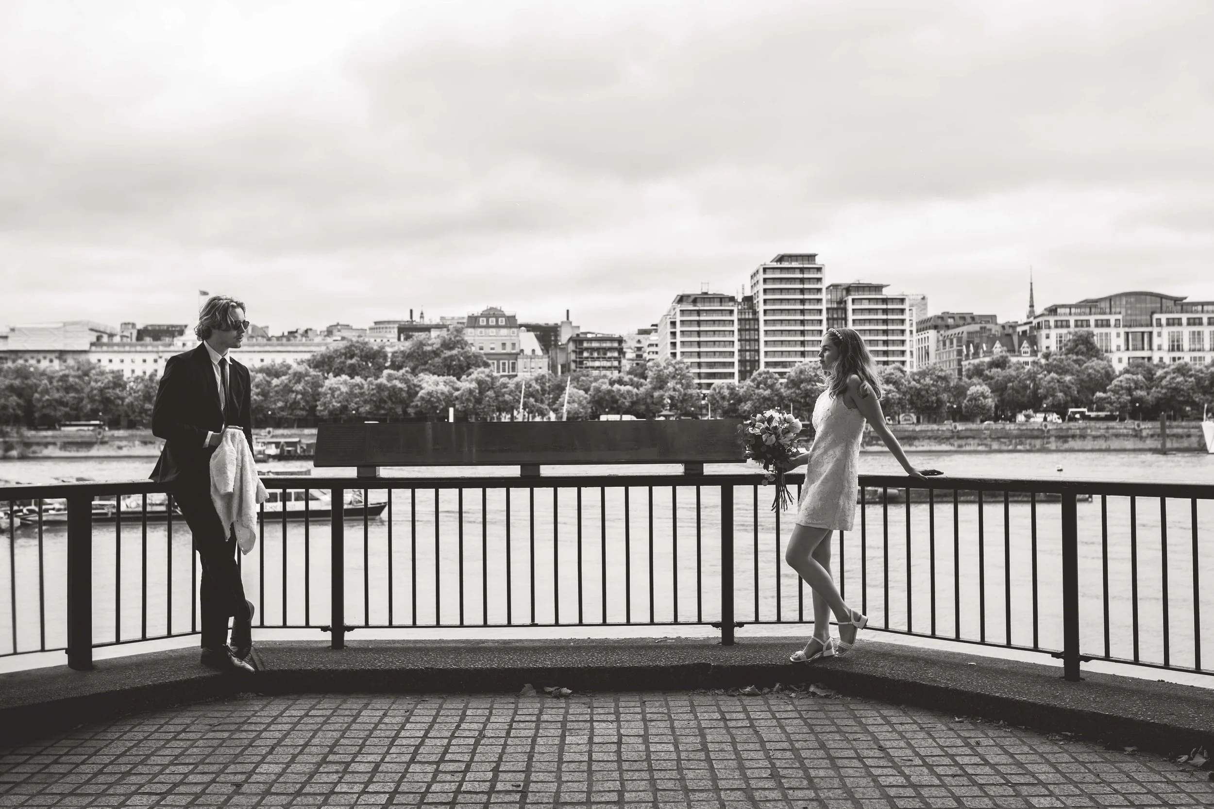 Black and white photo of a bride holding a bouquet, leaning against a railing by a river, with a groom standing nearby, cityscape with trees in the background.