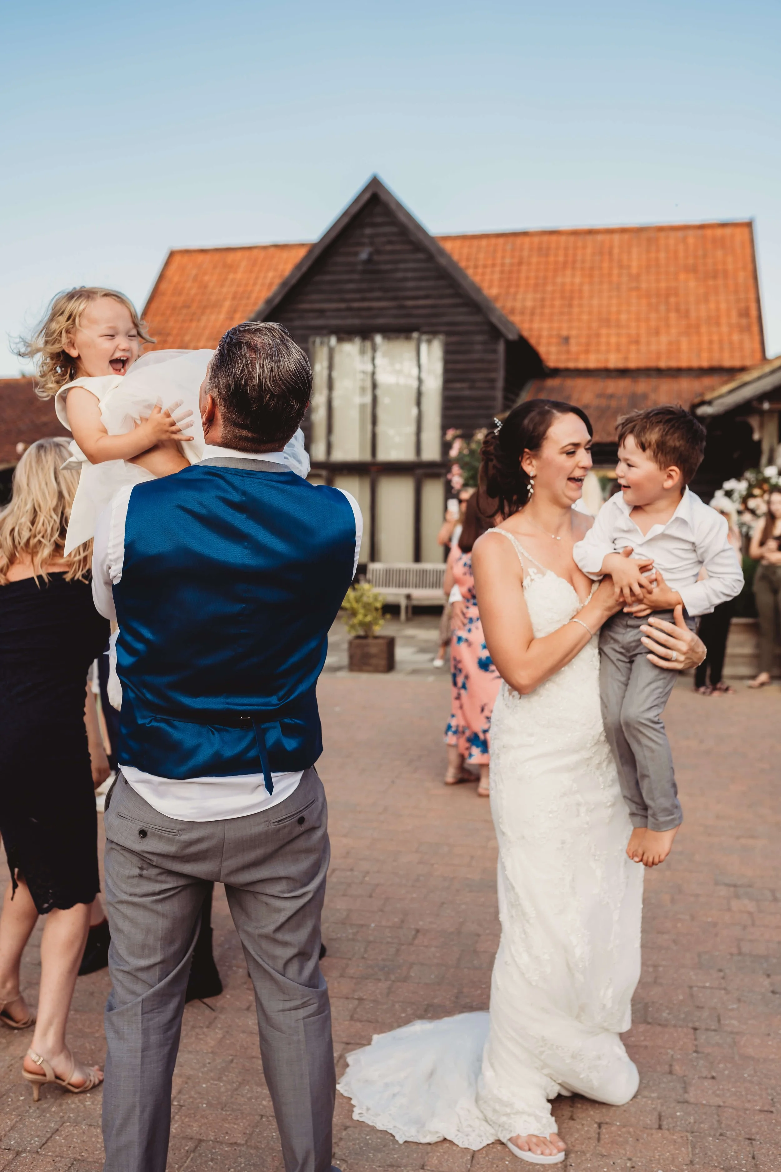 A wedding celebration outdoors with guests dancing and children playing, featuring a woman in a white wedding dress and a man holding a joyful girl.