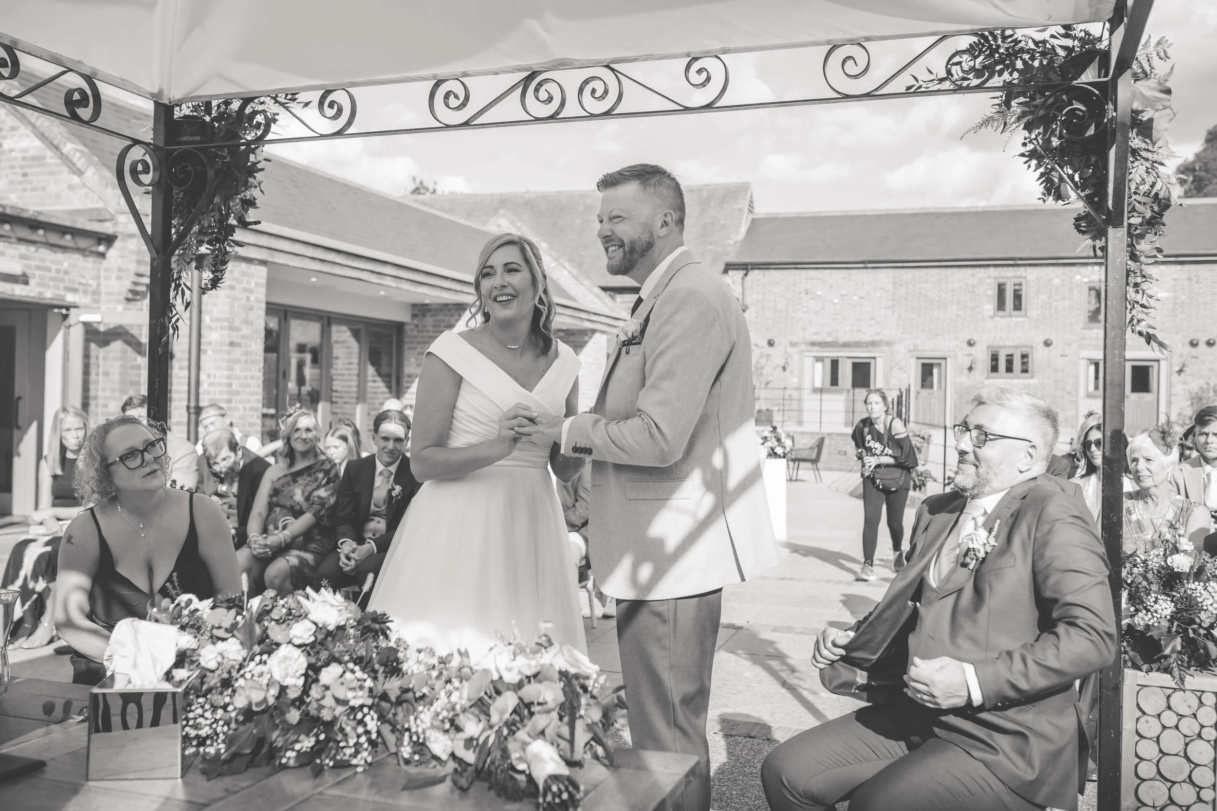 A black and white photo of an outdoor wedding ceremony, showing a bride and groom exchanging vows under a decorated canopy surrounded by seated guests. The bride is wearing a white wedding dress, and the groom is in a light-colored suit. There are bo