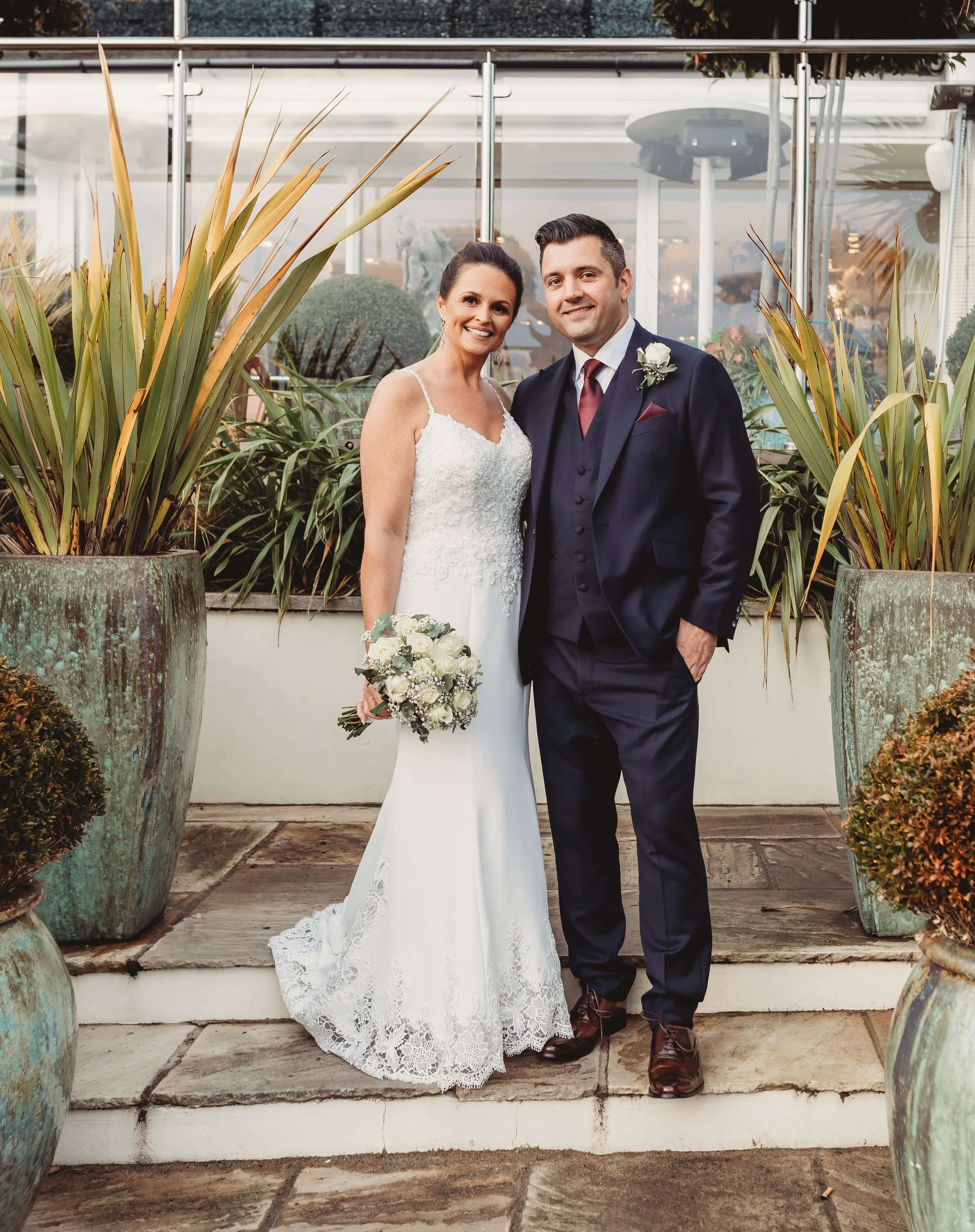 A bride in a white lace wedding dress and a groom in a dark suit, standing together outdoors on steps, surrounded by large potted plants and decorative shrubs.