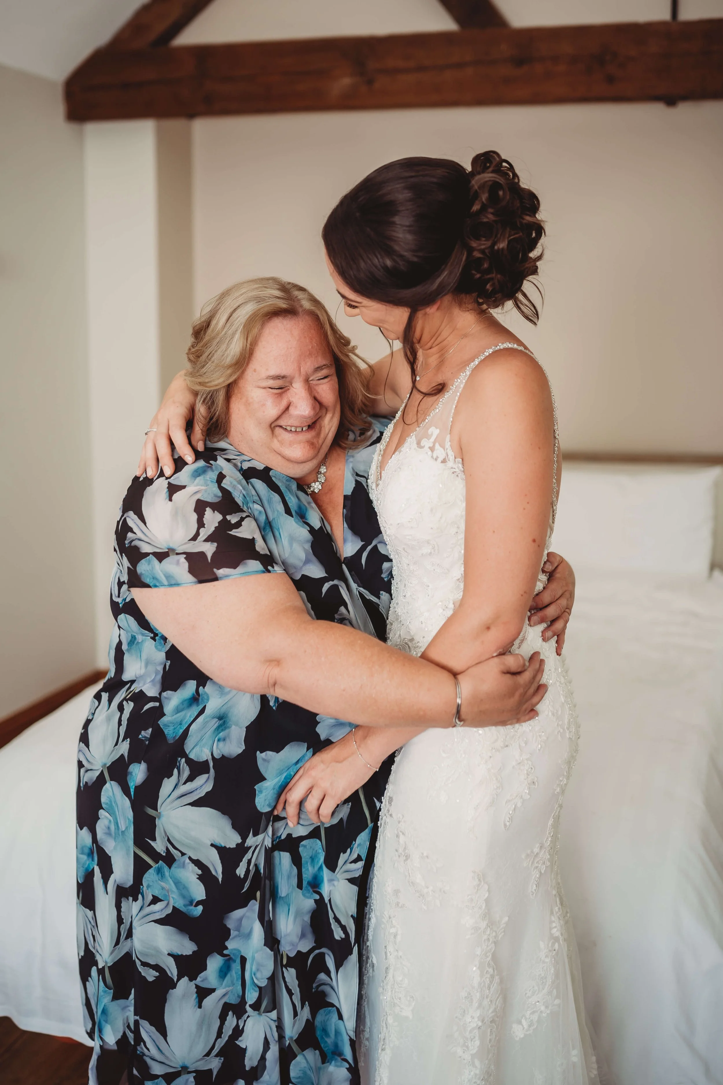 A joyful moment between a bride in a white wedding dress and an older woman in a floral dress, embracing and smiling in a bedroom.