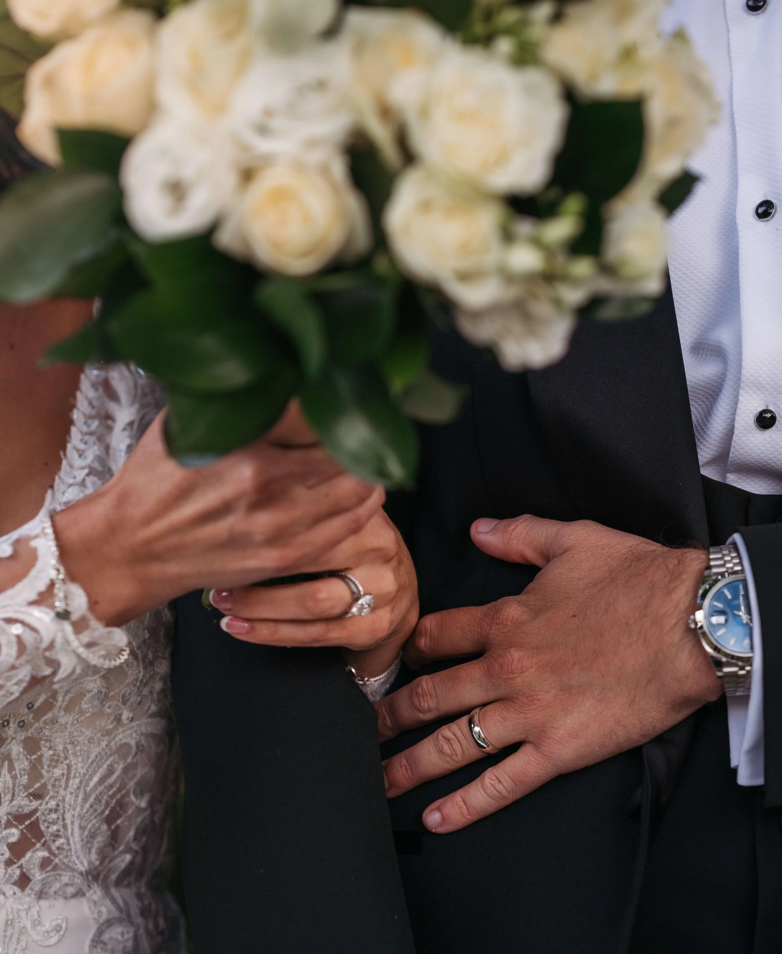 Close-up of a bride and groom's hands showing wedding rings with a bouquet and wedding attire visible.