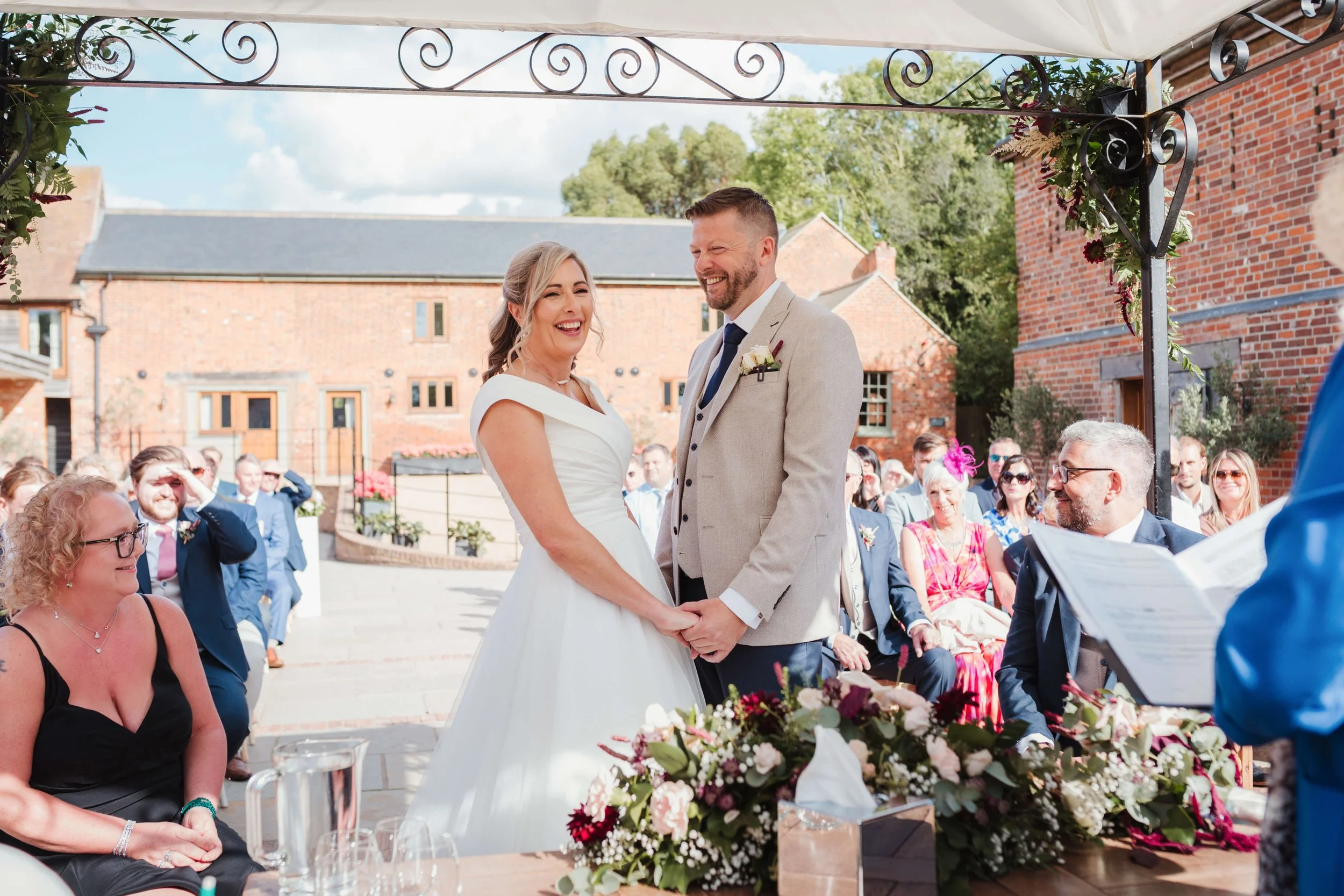 Bride and groom holding hands during wedding ceremony, smiling at each other, seated guests watching, outdoor setting with brick buildings and greenery, decorated arch, floral arrangement on table.