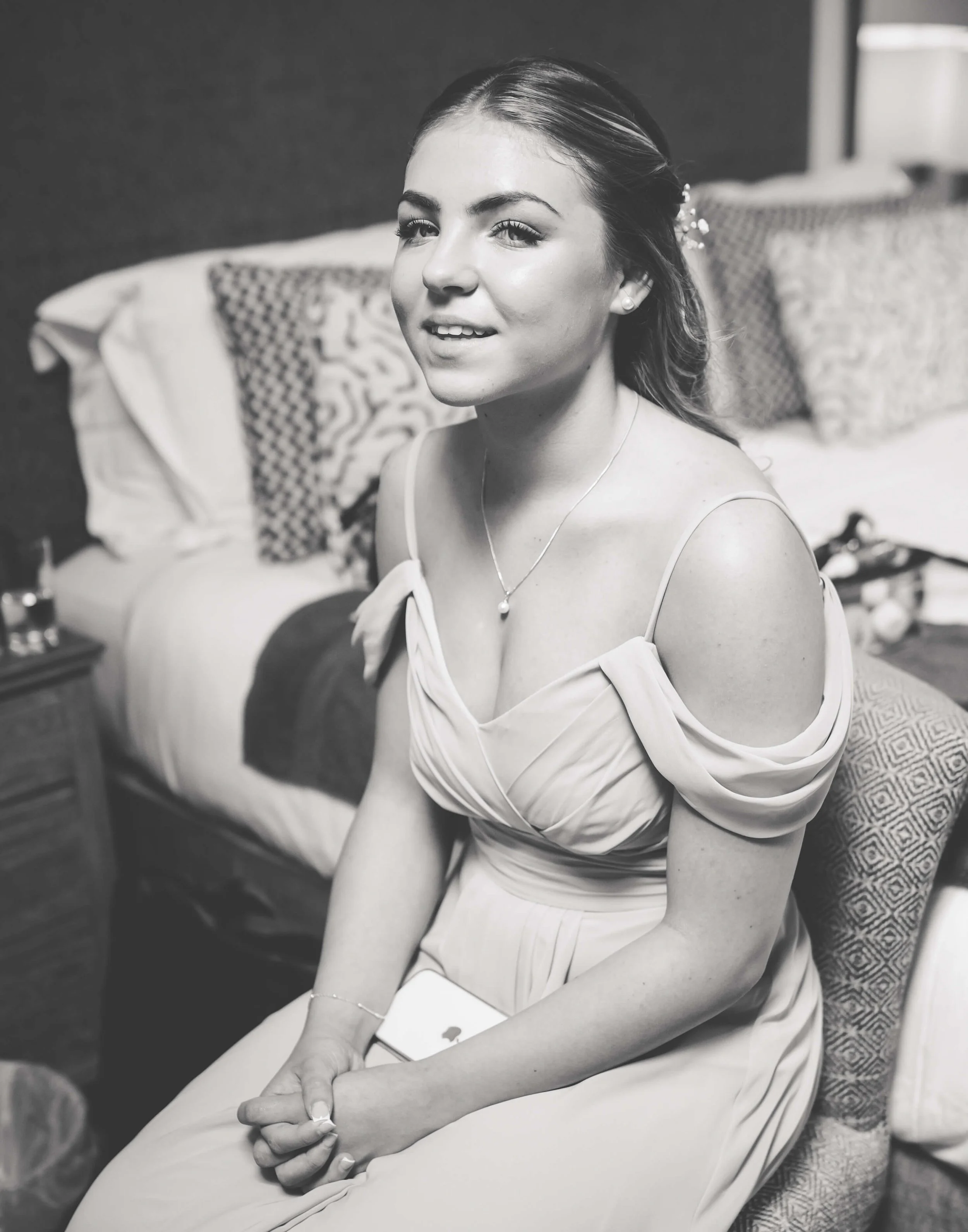 A young woman with dark hair styled back, wearing a light-colored dress with off-the-shoulder sleeves, sitting on a chair in a bedroom. She is smiling slightly and looking at the camera, with a necklace and earrings. The background features pillows a
