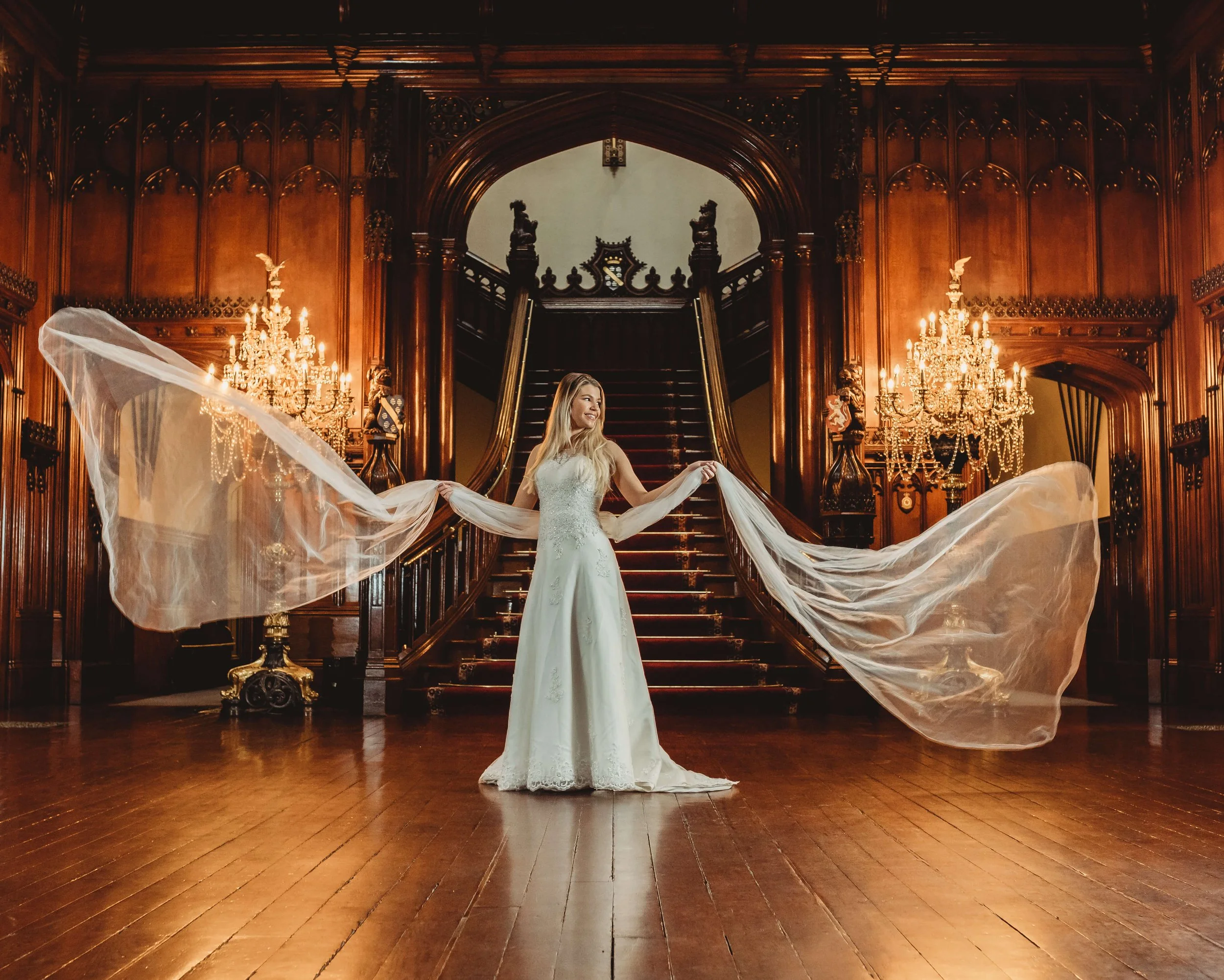 A woman in a white wedding dress stands in a grand, wood-paneled hall with ornate chandeliers, a staircase, and detailed woodwork, holding a sheer veil extended outward.