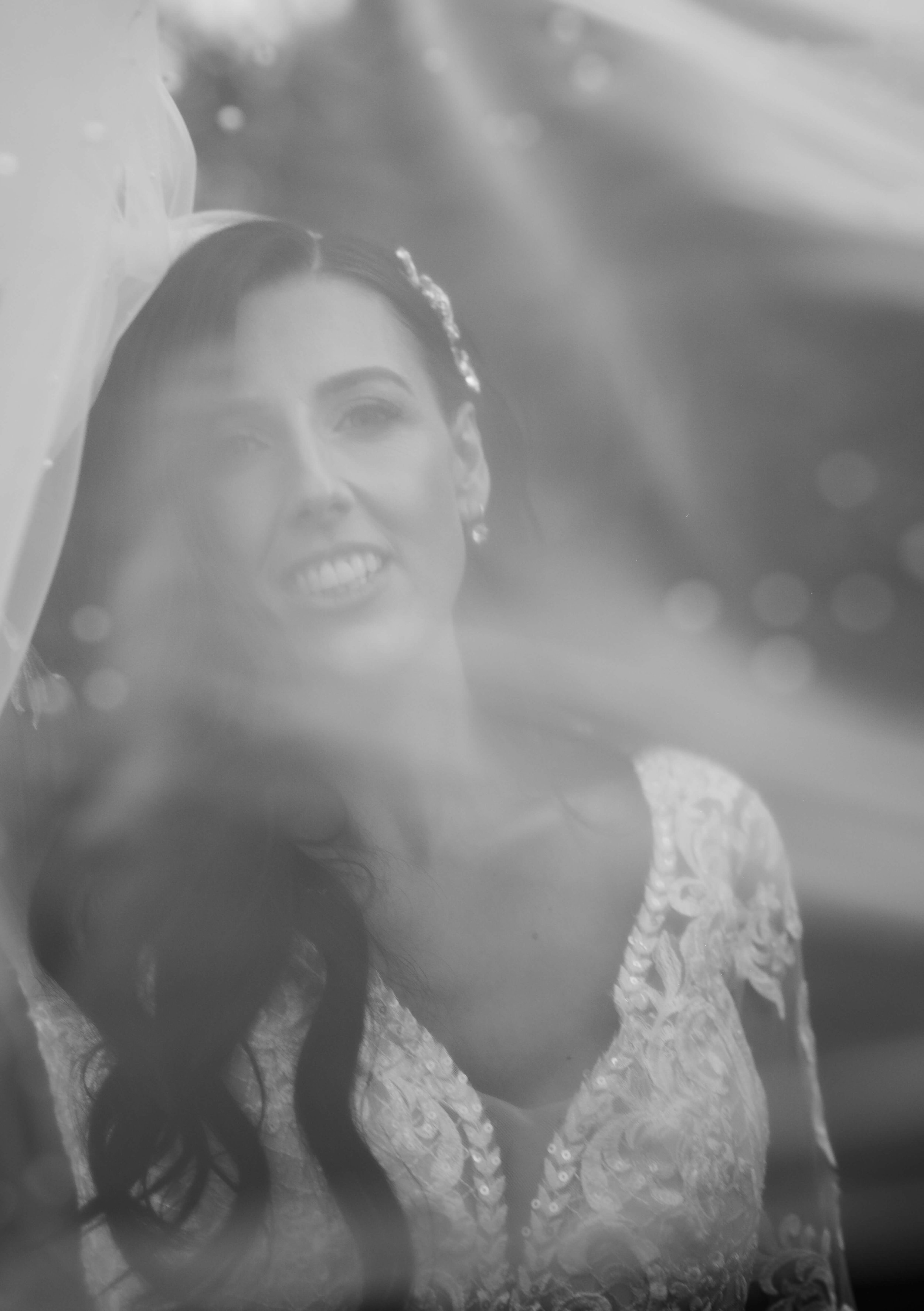Black and white photo of a woman with long dark hair, smiling slightly, wearing a lace dress and a hair accessory, seen through a slightly blurred glass or reflection.