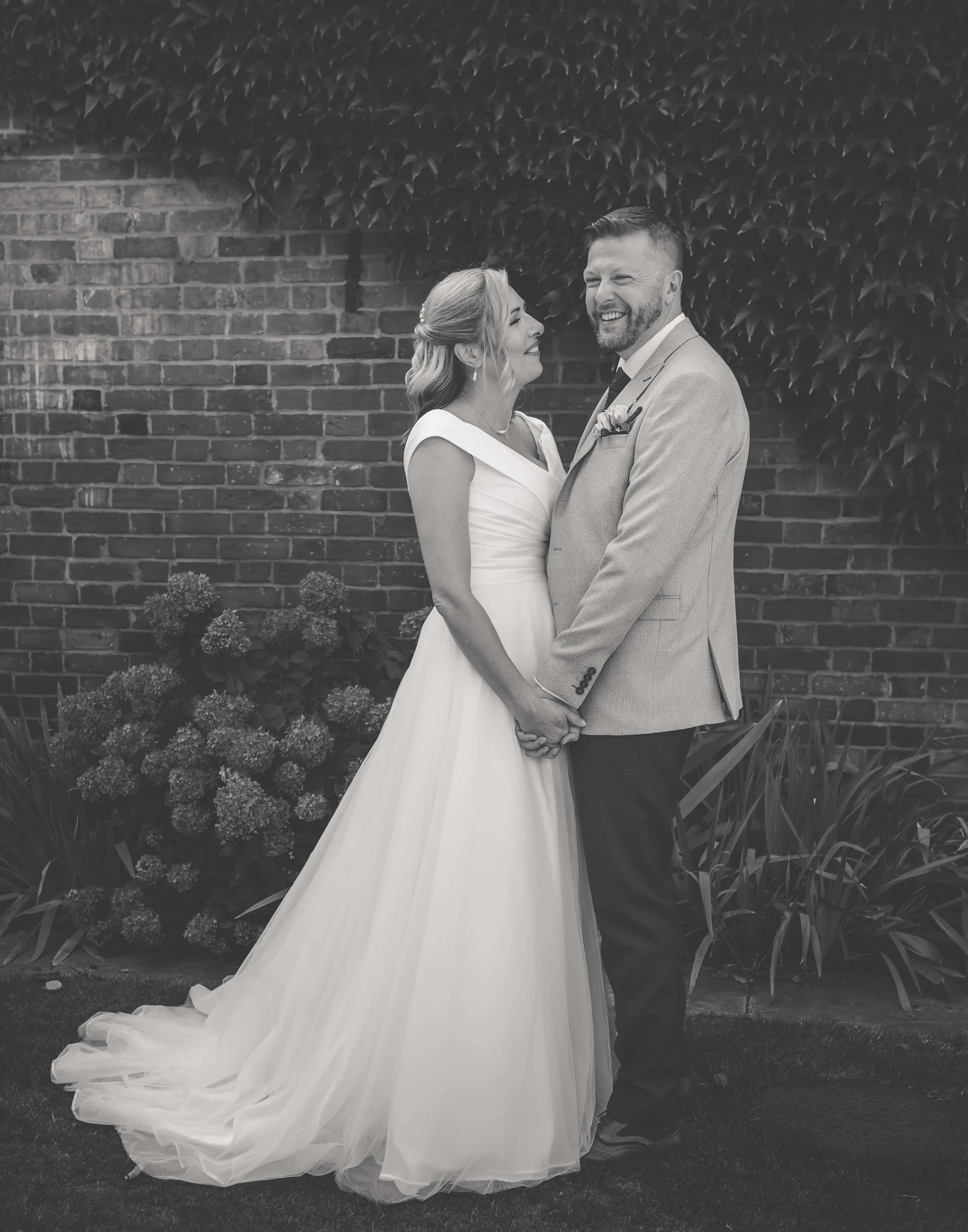 A black and white photo of a bride and groom holding hands, standing outdoors against a brick wall with ivy and flowers, smiling at each other.