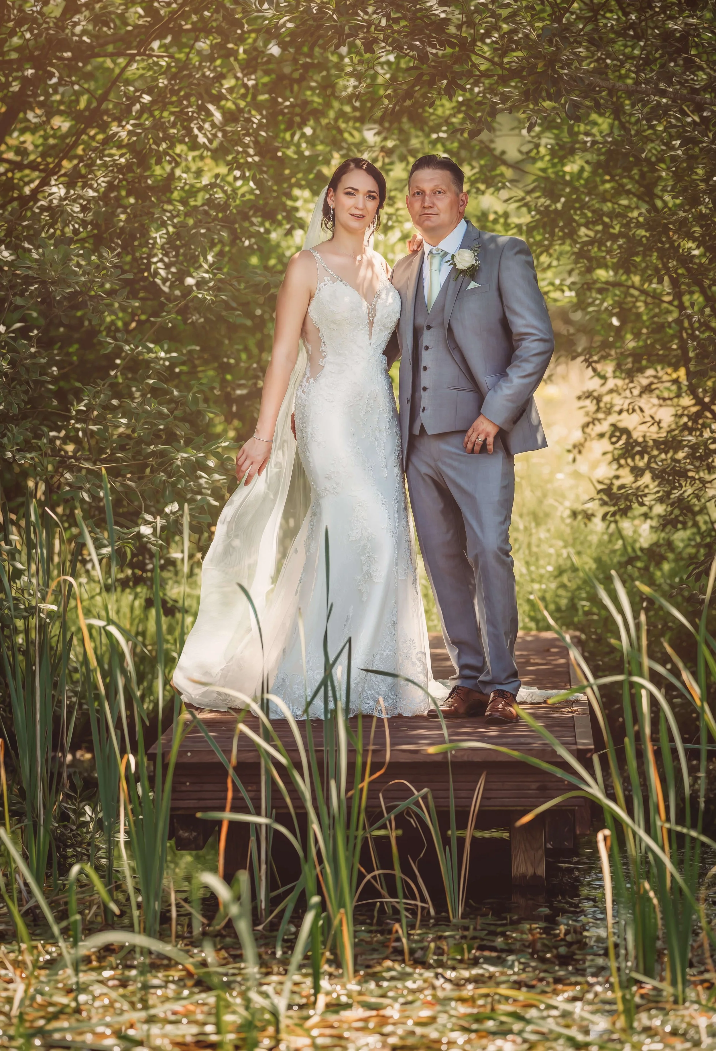 A bride and groom standing on a small wooden bridge surrounded by lush green trees and vegetation during daytime.