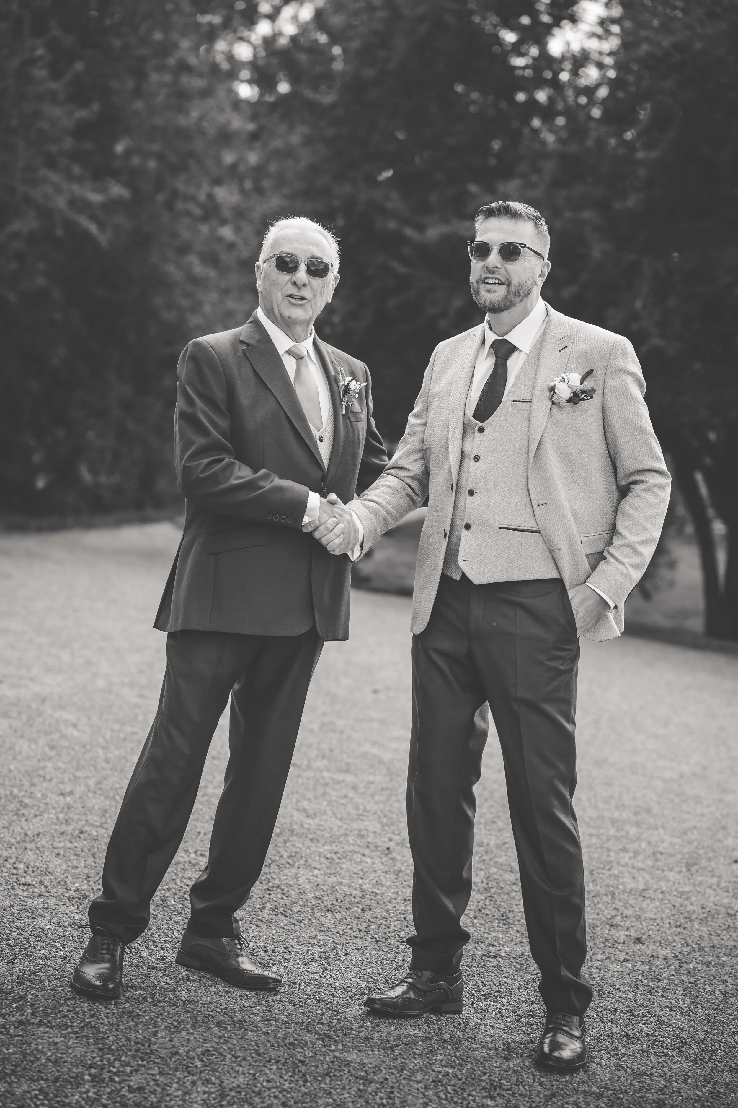 Two men in suits shaking hands outdoors, one older and one younger, both wearing sunglasses and boutonnières, possibly at a wedding or special event.