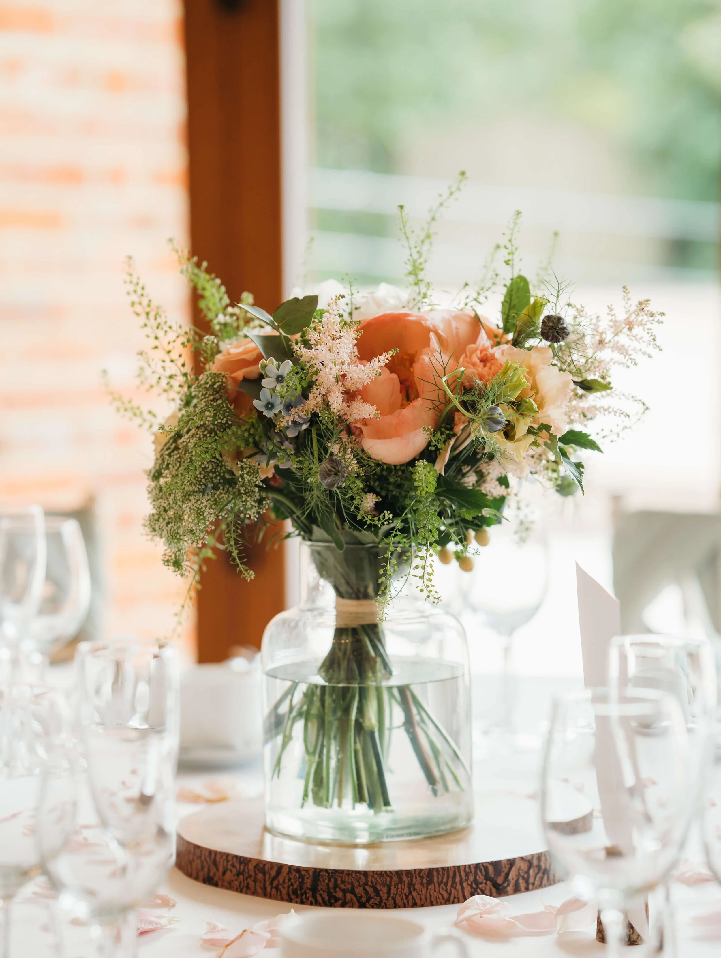 A floral centerpiece with peach, white, and green flowers in a clear glass vase on a round wooden table, surrounded by wine glasses and pink flower petals, indoors near a window