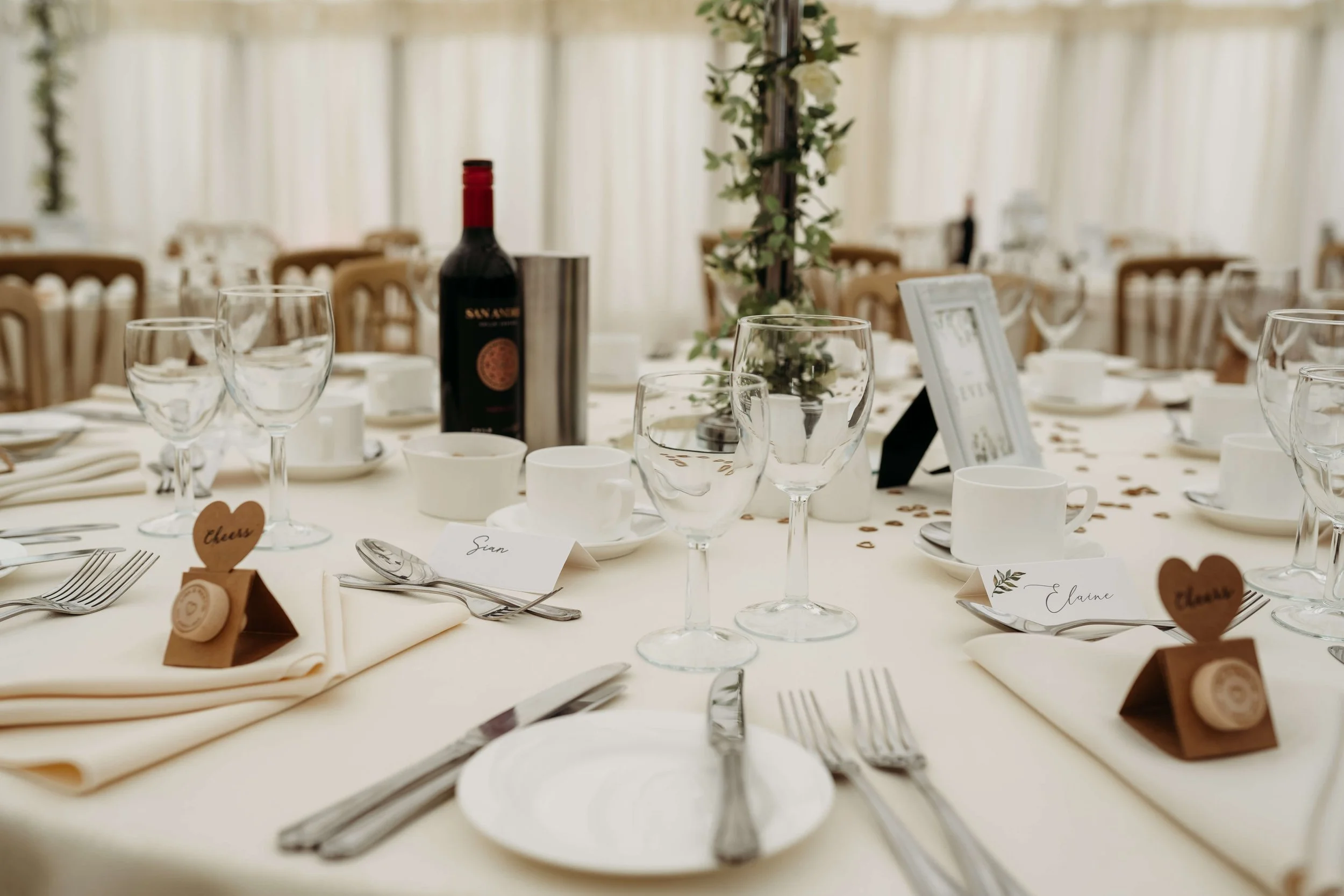 A banquet table set for a wedding reception with white tablecloths, wine and water glasses, teacups, silverware,place cards with guest names, a bottle of red wine, and a floral centerpiece.