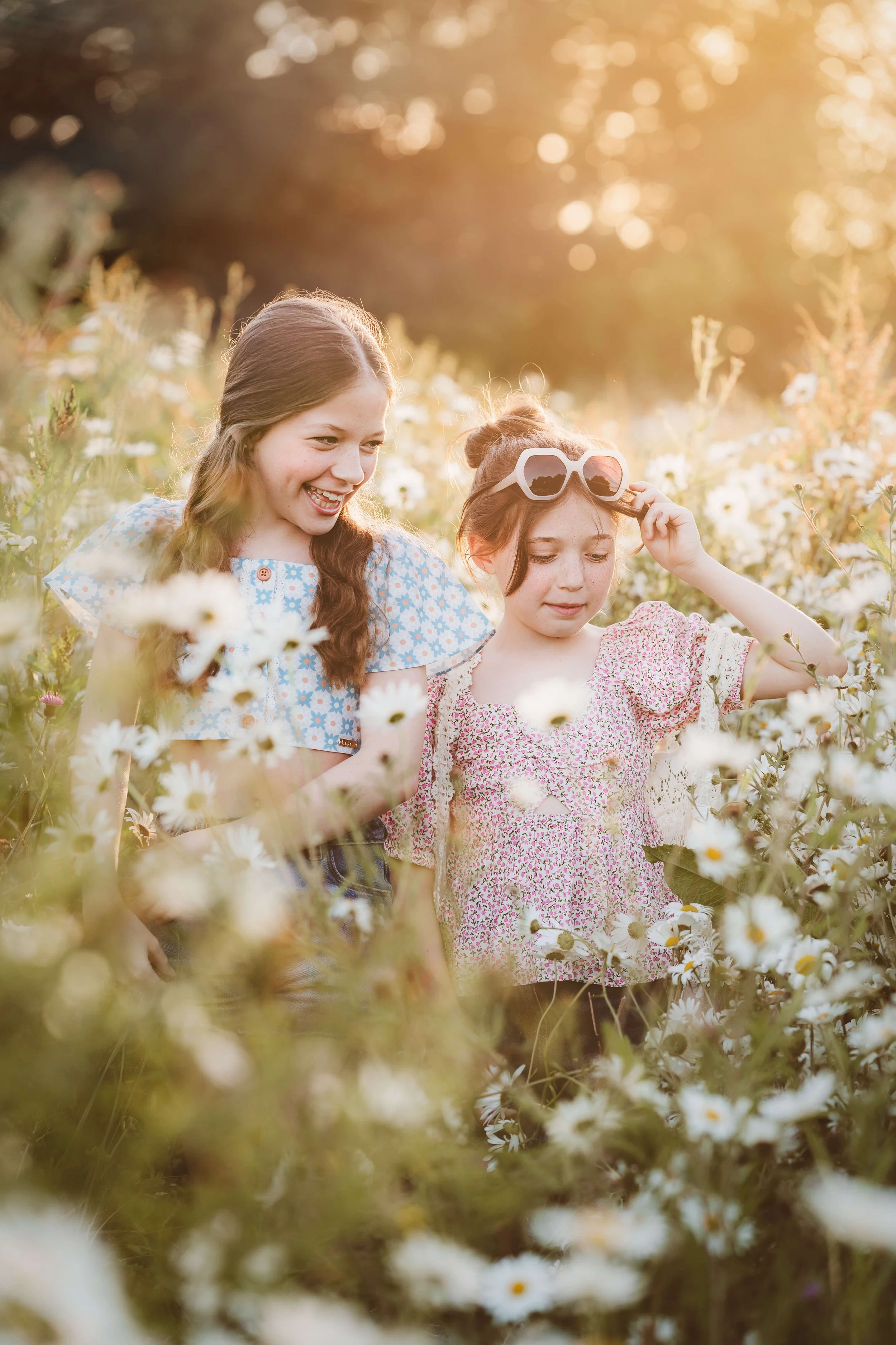 Two young girls enjoying a daisy field during sunset, one with sunglasses on her head, both smiling.