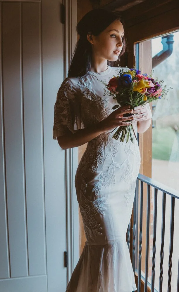 A woman in a white floral dress holding a colorful bouquet of flowers, standing near a window and looking outside.