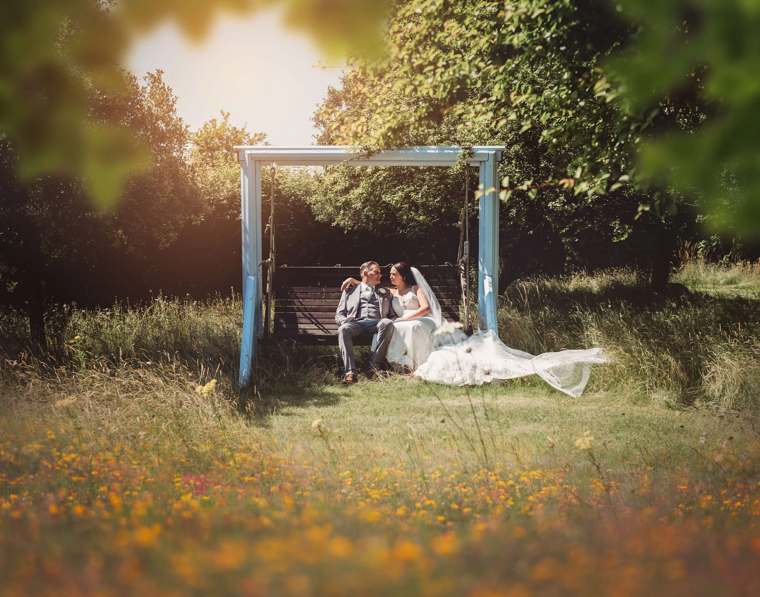 A bride and groom sitting close on a porch swing in a grassy area, surrounded by trees and flowers, during golden hour.