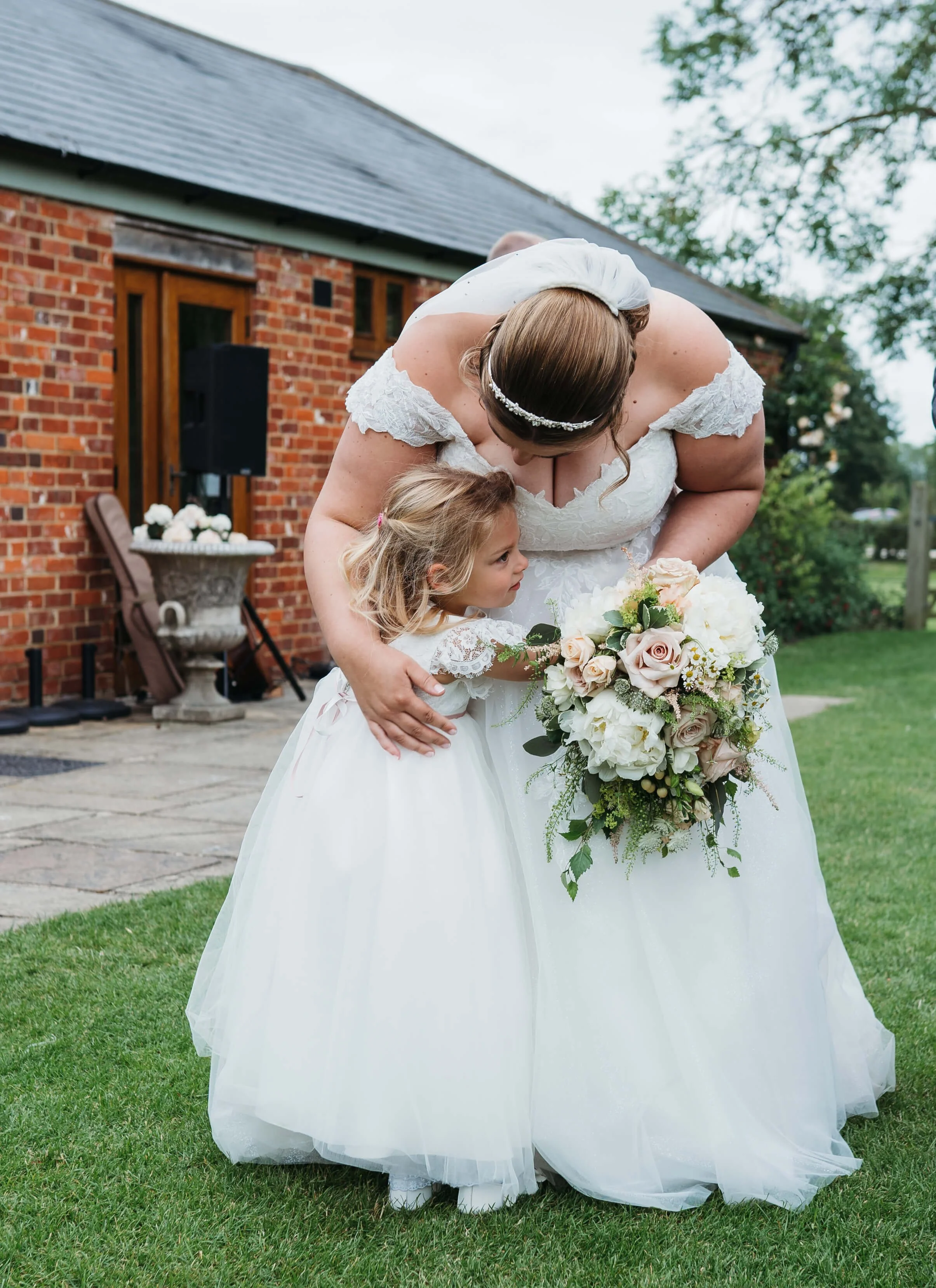 A bride in a white wedding dress with lace shoulders is hugging a young girl in a white dress with lace details outside near a brick building, holding a bouquet of flowers.