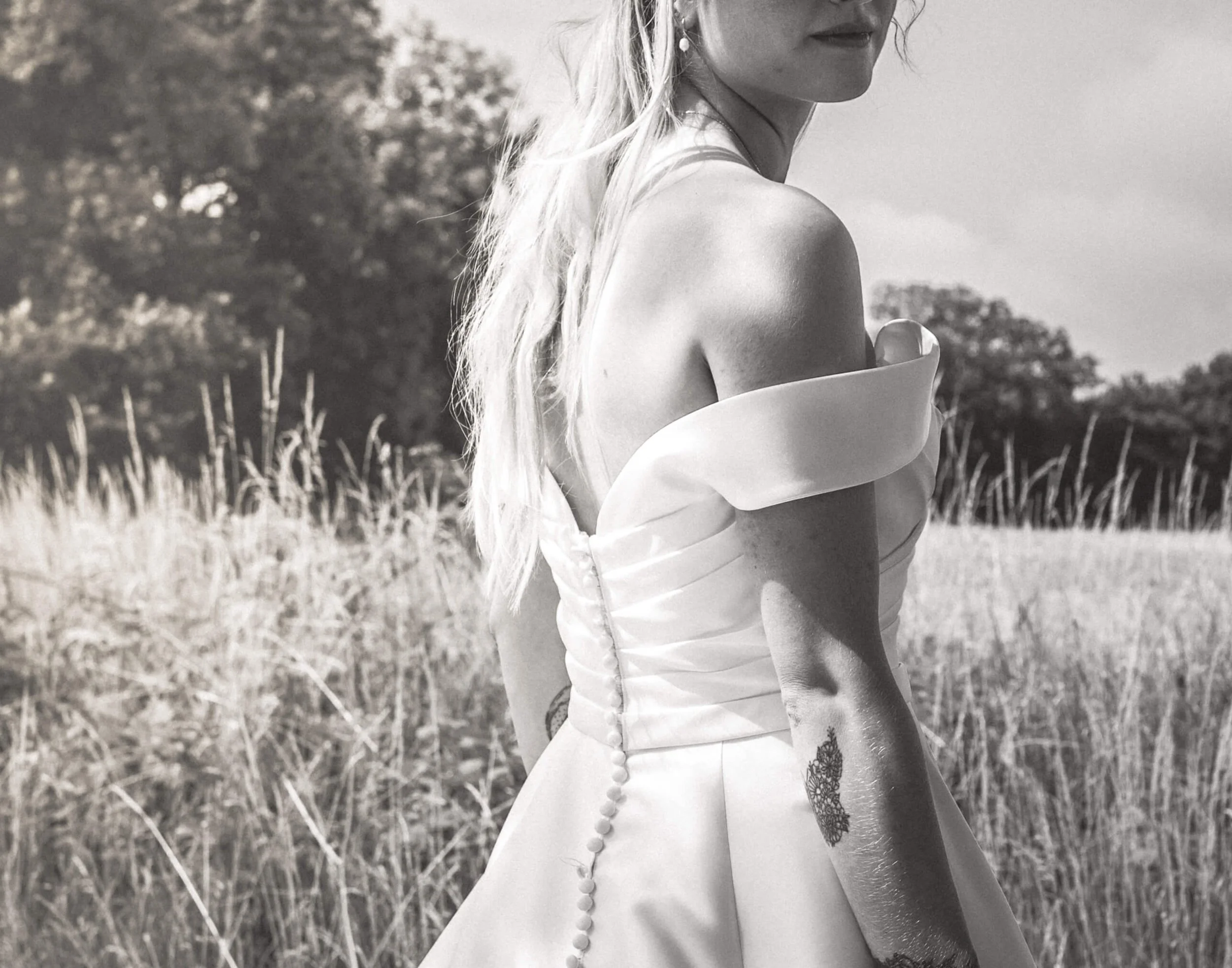 A black and white photo of a woman in an off-shoulder dress standing in a field of grass with trees in the background.