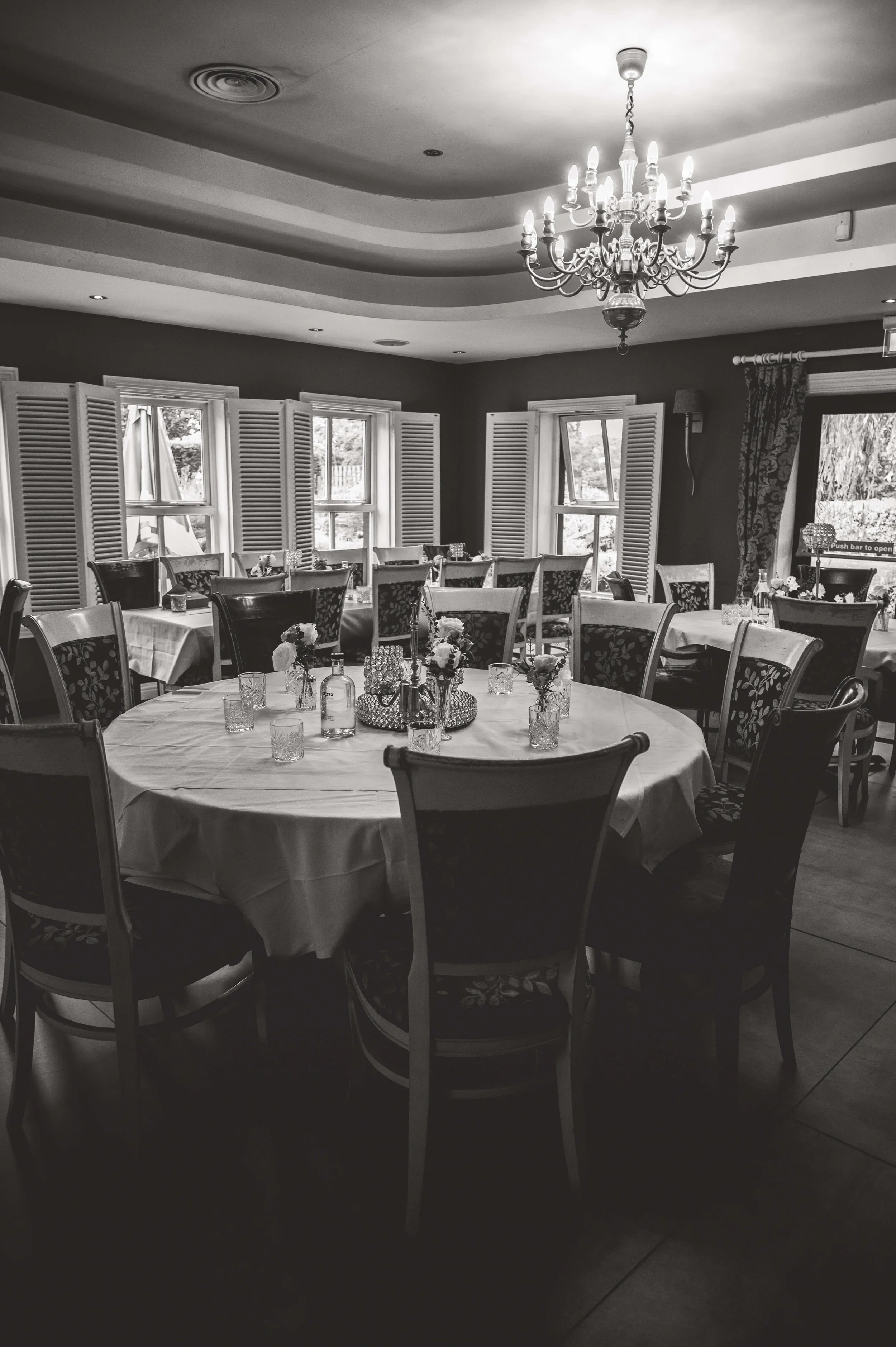 A decorated dining room with round tables covered in white tablecloths, surrounded by chairs with floral and plain upholstery, topped with flower centerpieces and glassware. Windows with shutters allow natural light to fill the room, and a chandelier