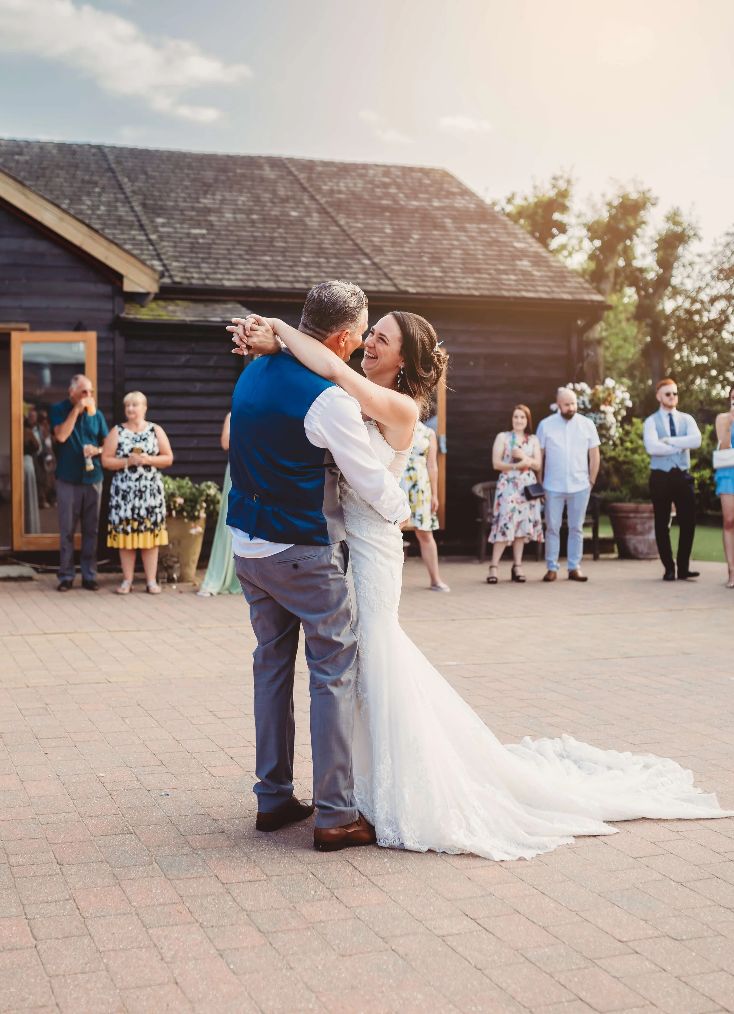 A bride and groom dance closely outside at sunset, surrounded by friends and family, with a rustic dark wooden building in the background.