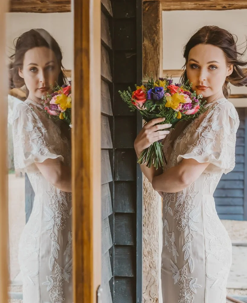 A woman in a white lace dress holding a colorful bouquet of flowers, standing next to a mirror reflecting her face and upper body.