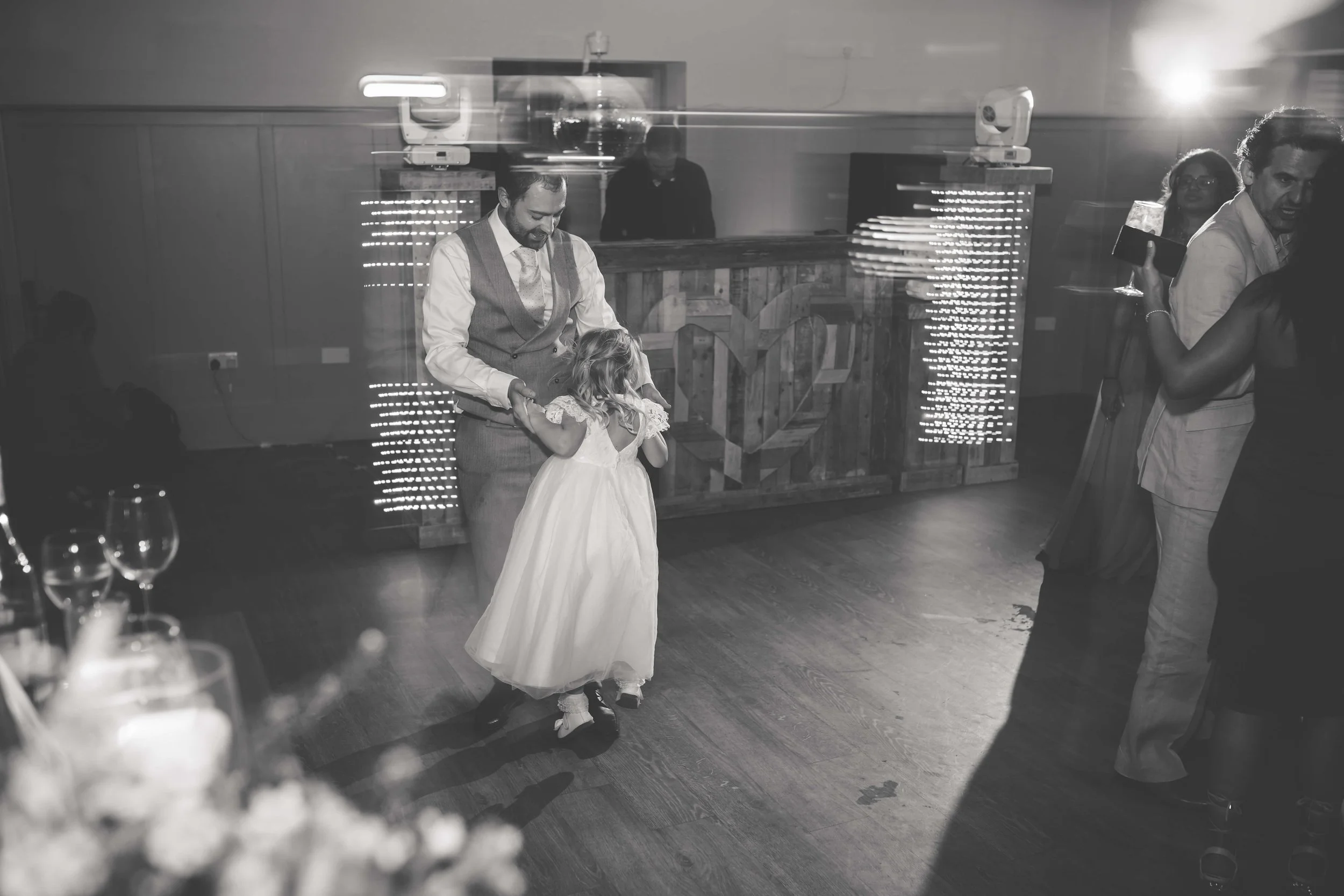 A man in a vest and tie dancing with a young girl in a white dress at a wedding reception, with a DJ booth in the background and other guests dancing.