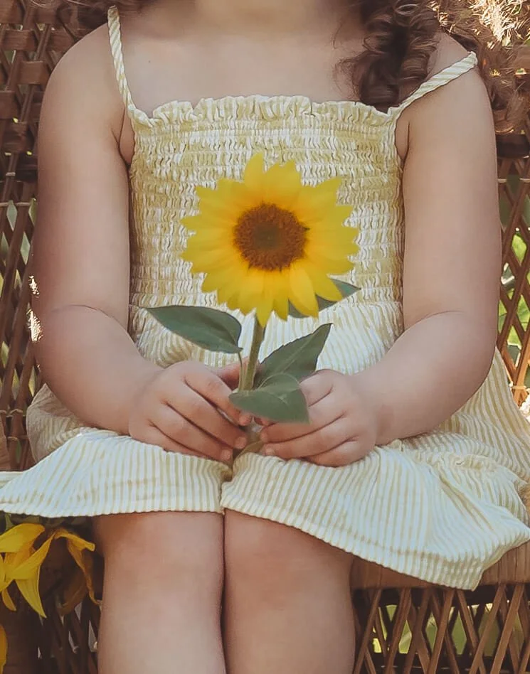 A young girl in a yellow and white striped dress sitting on a wooden chair, holding a large sunflower with green leaves.