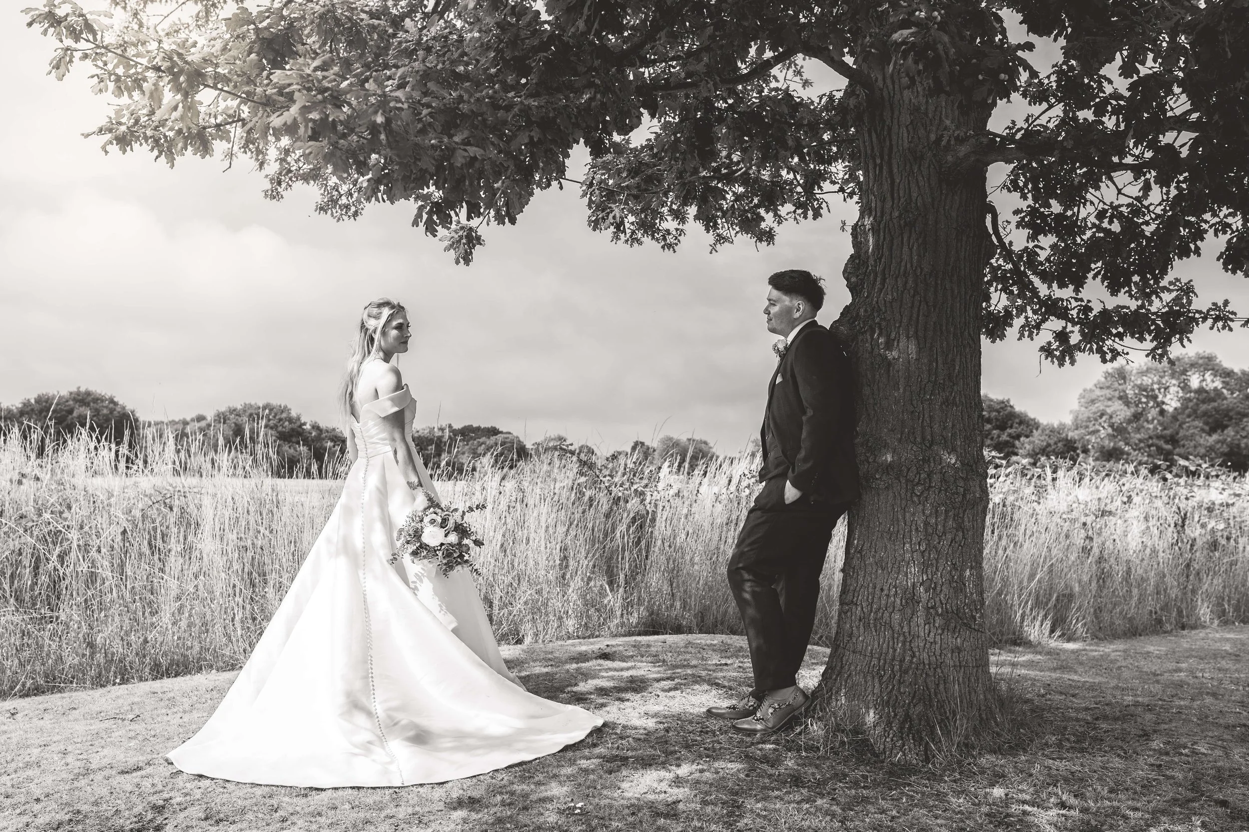 Black and white photo of a bride and groom outdoors near a large tree. The bride wears a long wedding gown and holds a bouquet, while the groom, dressed in a black tuxedo, leans against the tree looking at her.