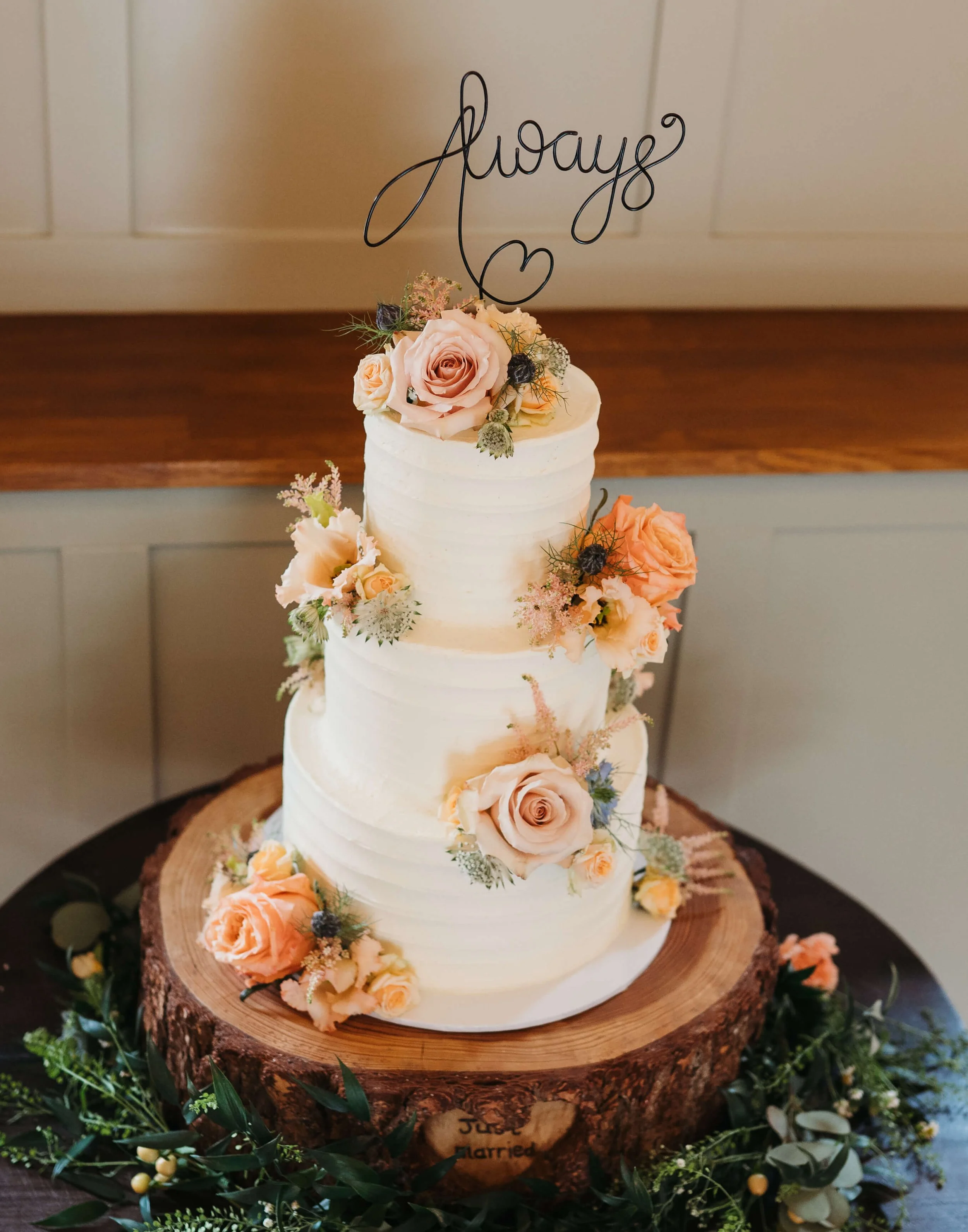 A three-tiered wedding cake decorated with pastel peach roses and greenery, topped with a wire cake topper spelling 'Always' with a heart. The cake is on a wooden slice with greenery around it.