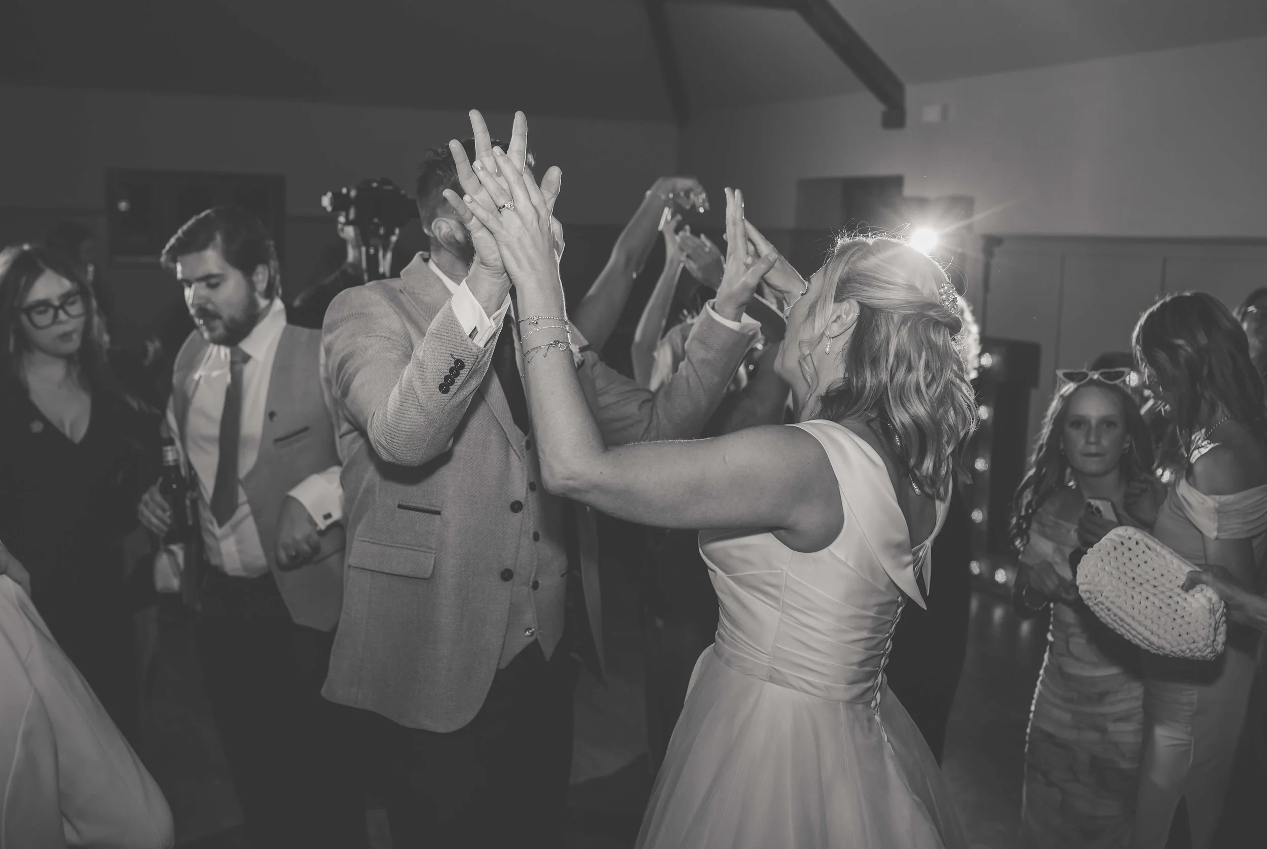 People dancing at a wedding reception, center focus on a couple in formal attire, with the woman in a white dress and the man in a light-colored suit, sharing a dance.