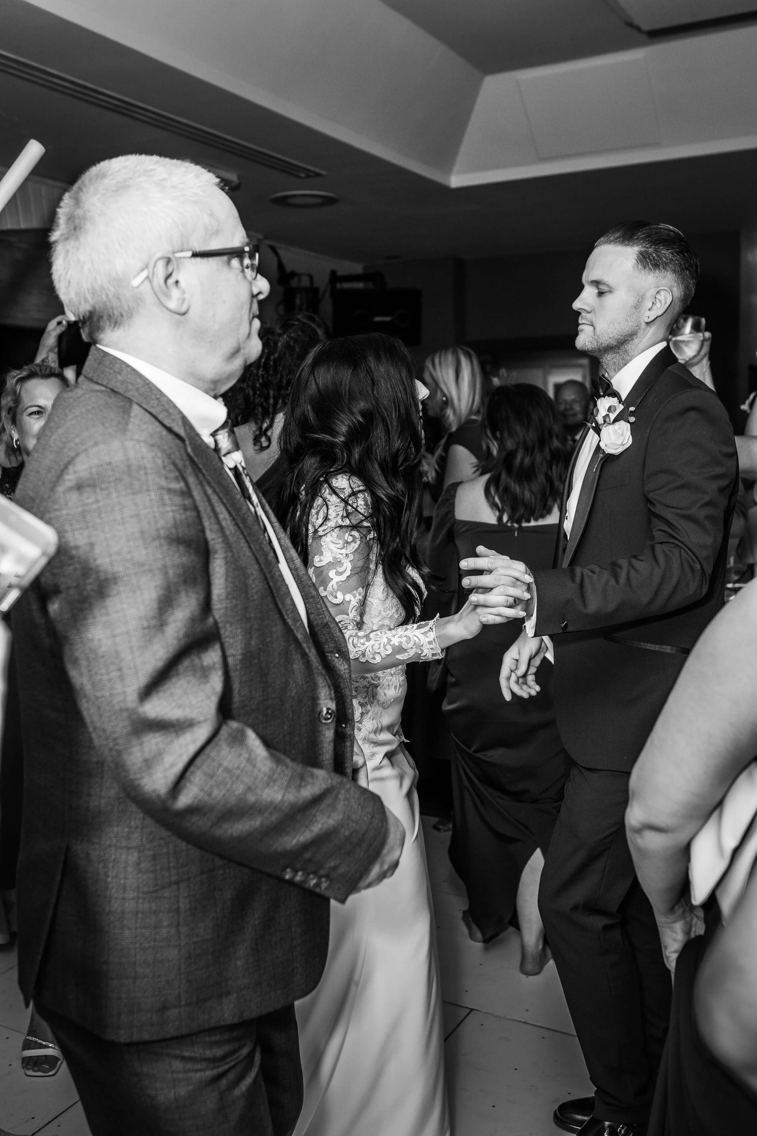 A black and white photo of a wedding reception; a groom in a tuxedo and bow tie and a bride in a lace gown are dancing together, surrounded by guests.