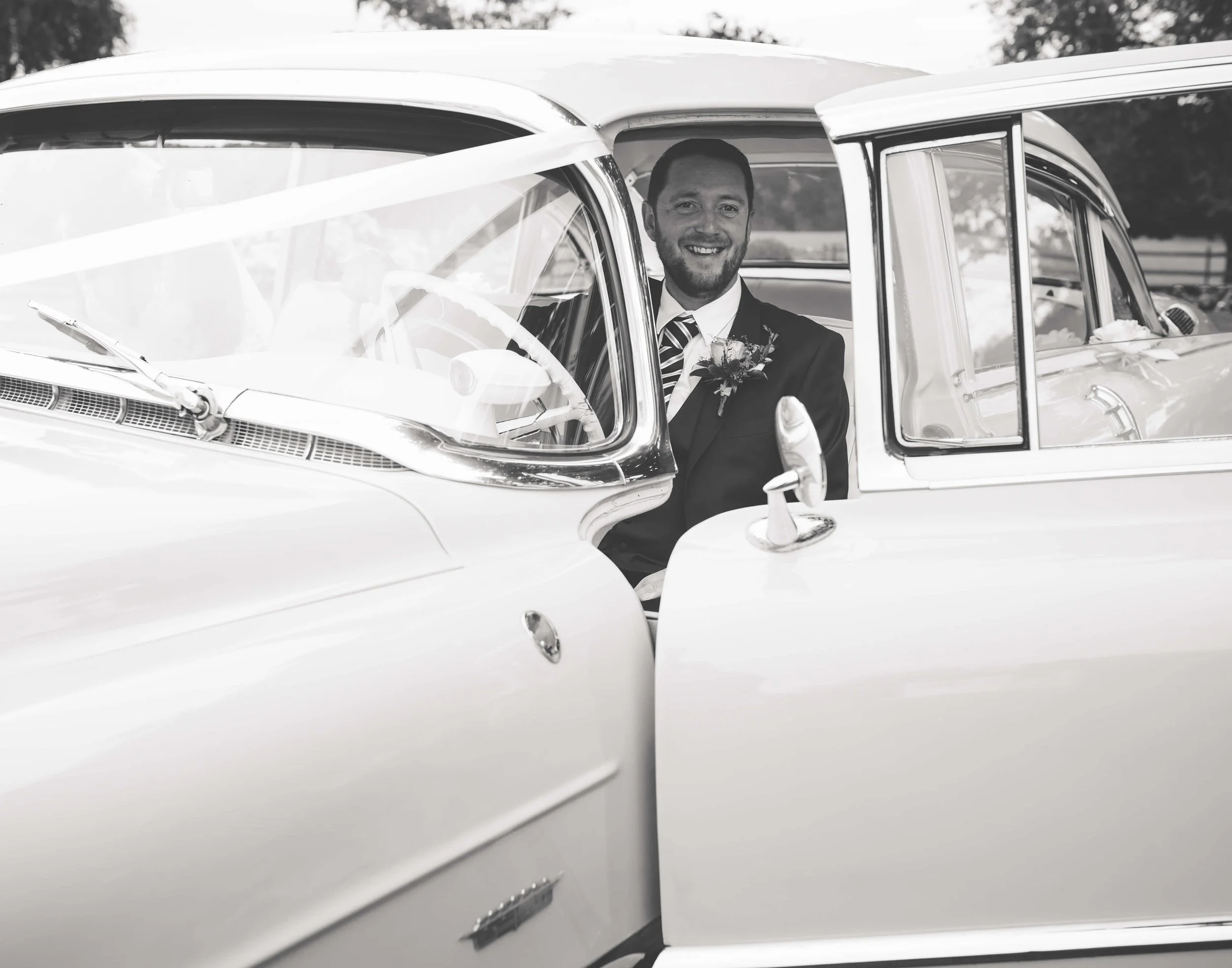 A man in a suit and tie sitting inside a vintage car, smiling, with trees in the background.