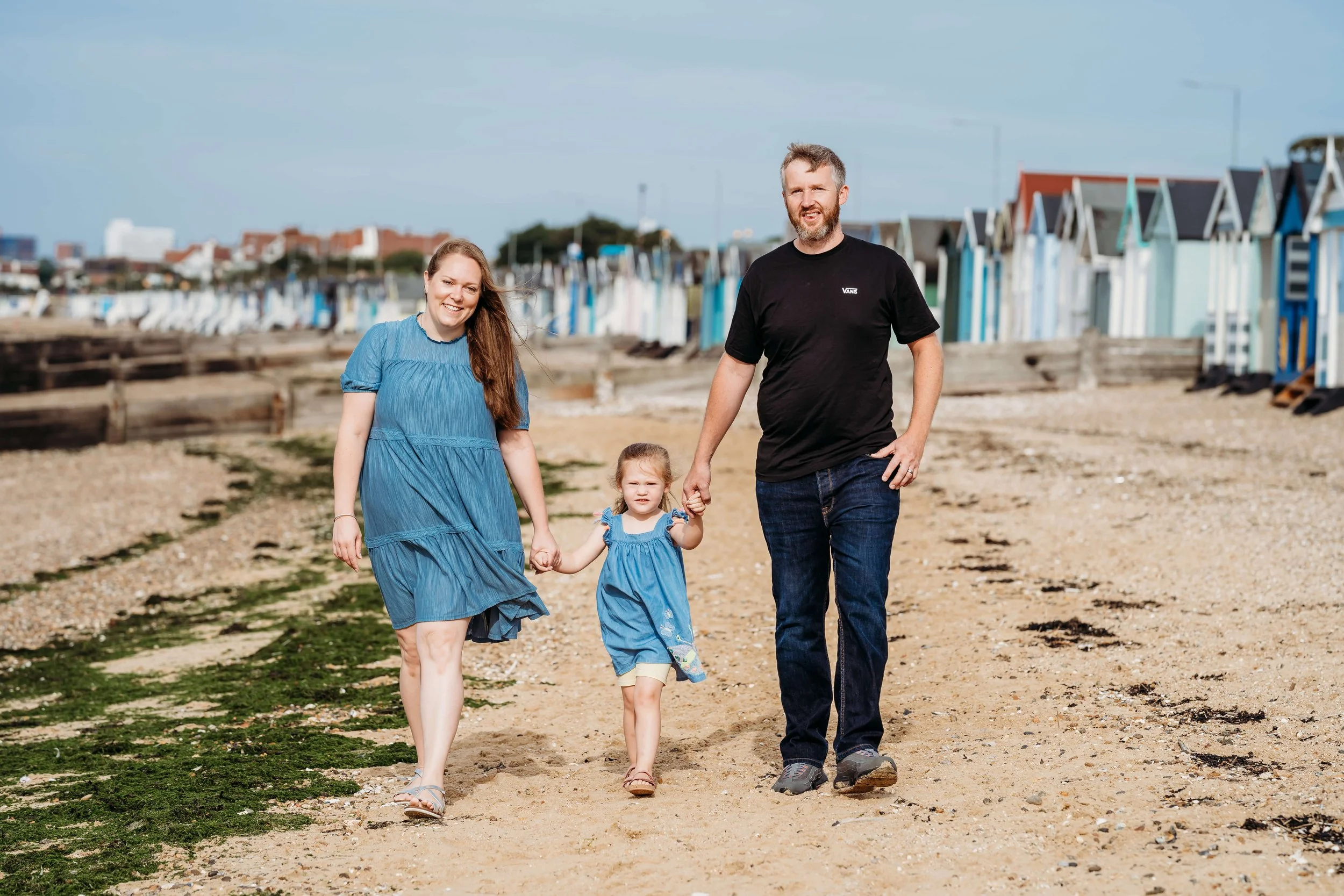 A family of three walking on a sandy beach, holding hands. The mother and daughter are smiling, while the father looks ahead. Beach huts line the background under a clear sky.