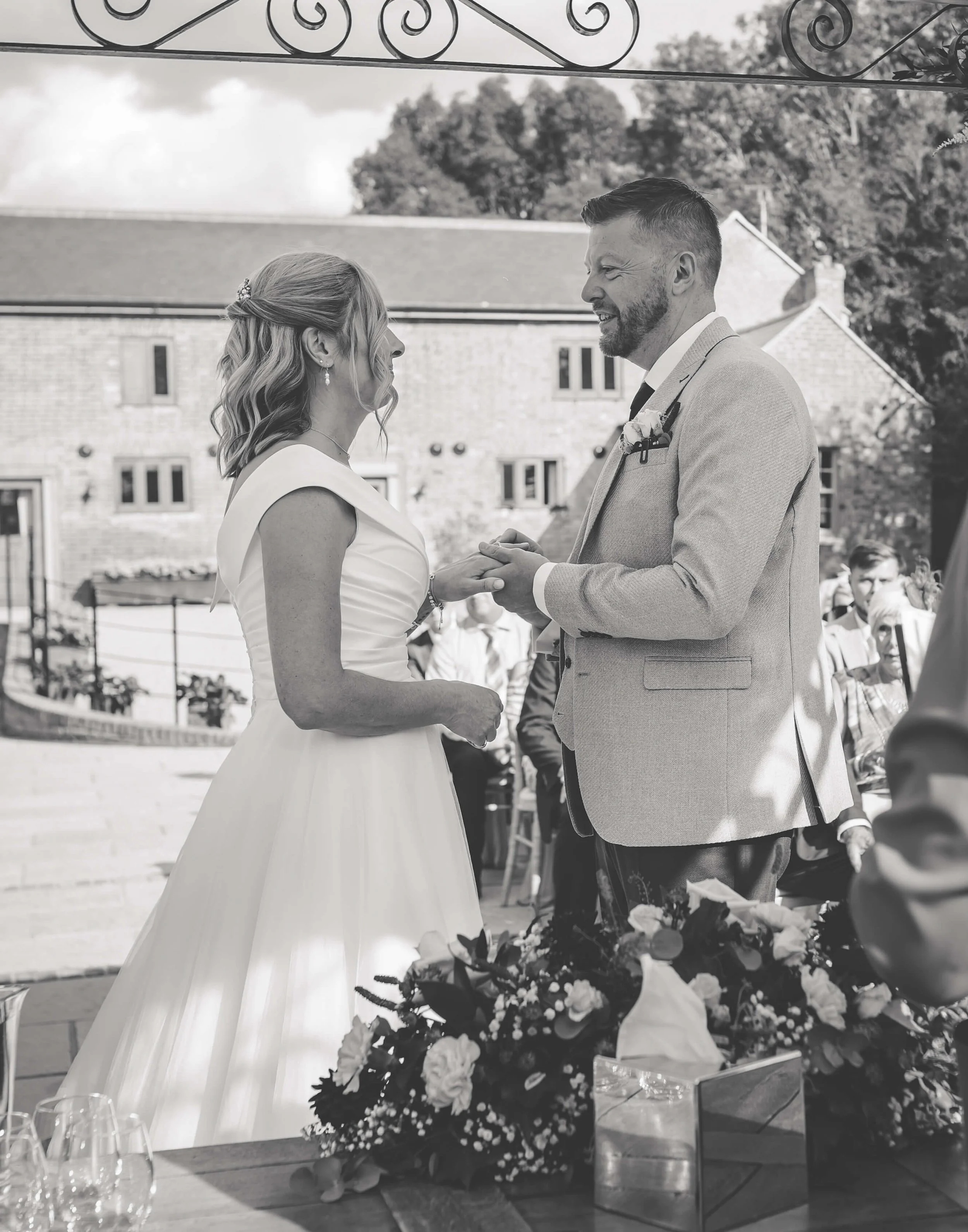 A black and white photo of a bride and groom exchanging wedding vows outdoors. The bride is wearing a sleeveless wedding dress, and the groom is dressed in a light-colored suit. They are holding hands and gazing at each other, with guests in the back