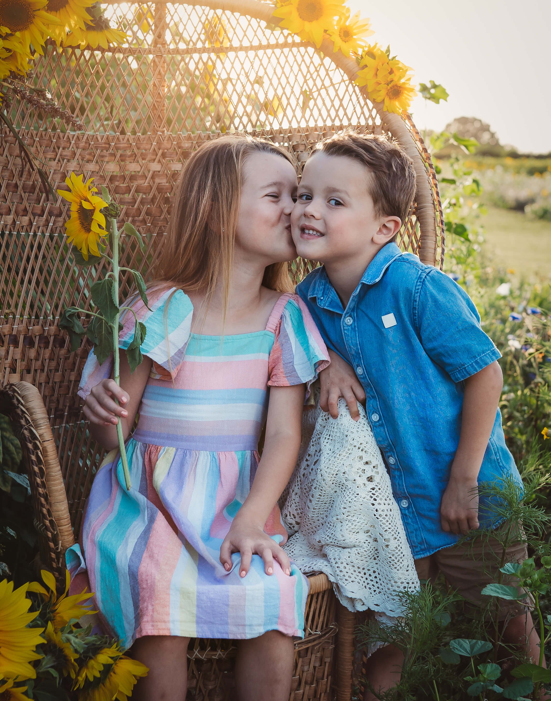 A young girl with red hair, wearing a colorful striped dress, kissing a young boy on the cheek. The boy, with dark hair, is wearing a blue denim shirt and beige shorts. They are sitting on a wicker chair surrounded by sunflowers in a garden during go