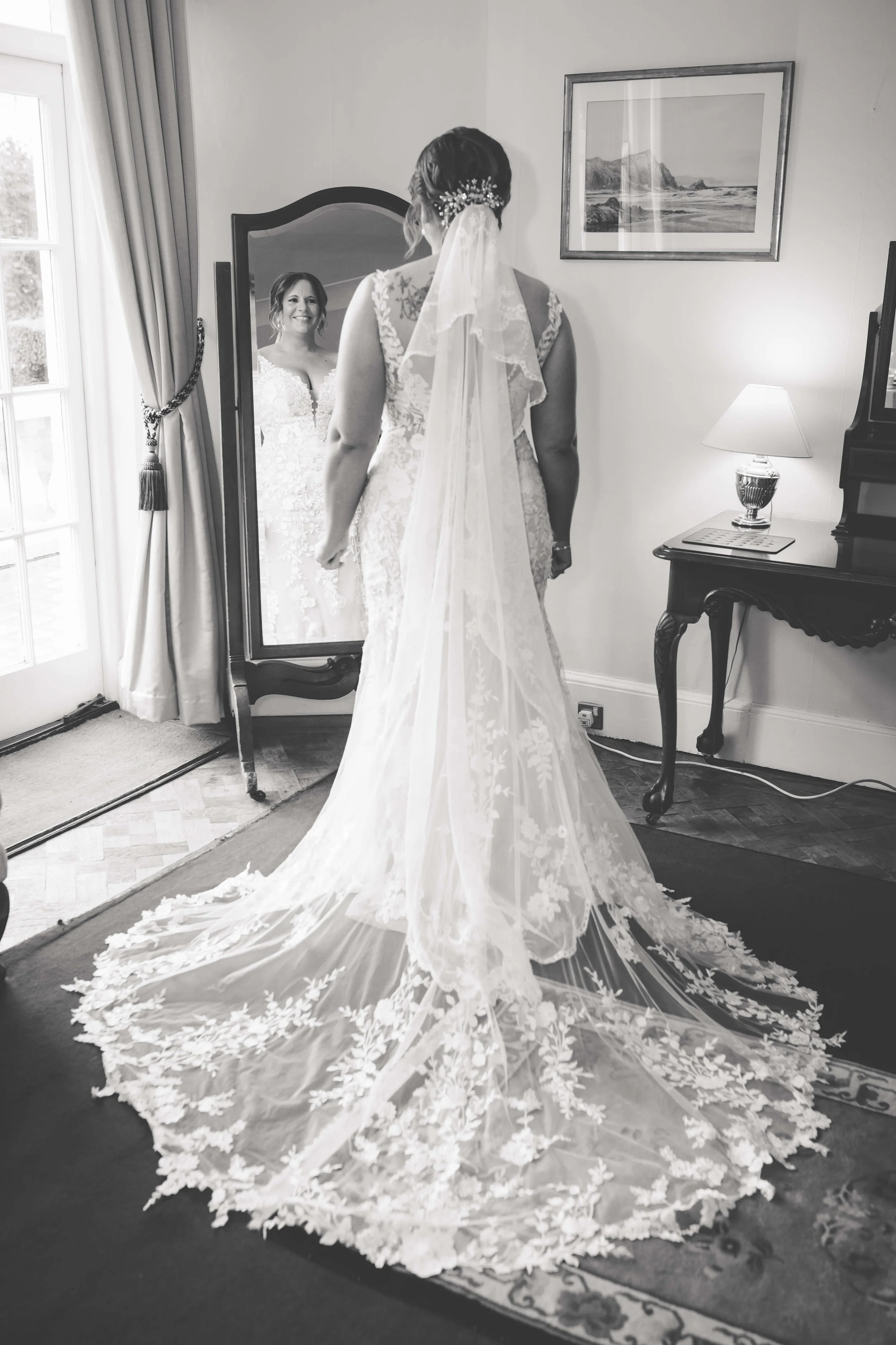 Bride in wedding dress looking at herself in a full-length mirror, with a wedding dress train decorated with floral lace patterns, in a room with a window, curtain, picture frame, and a table with a lamp.