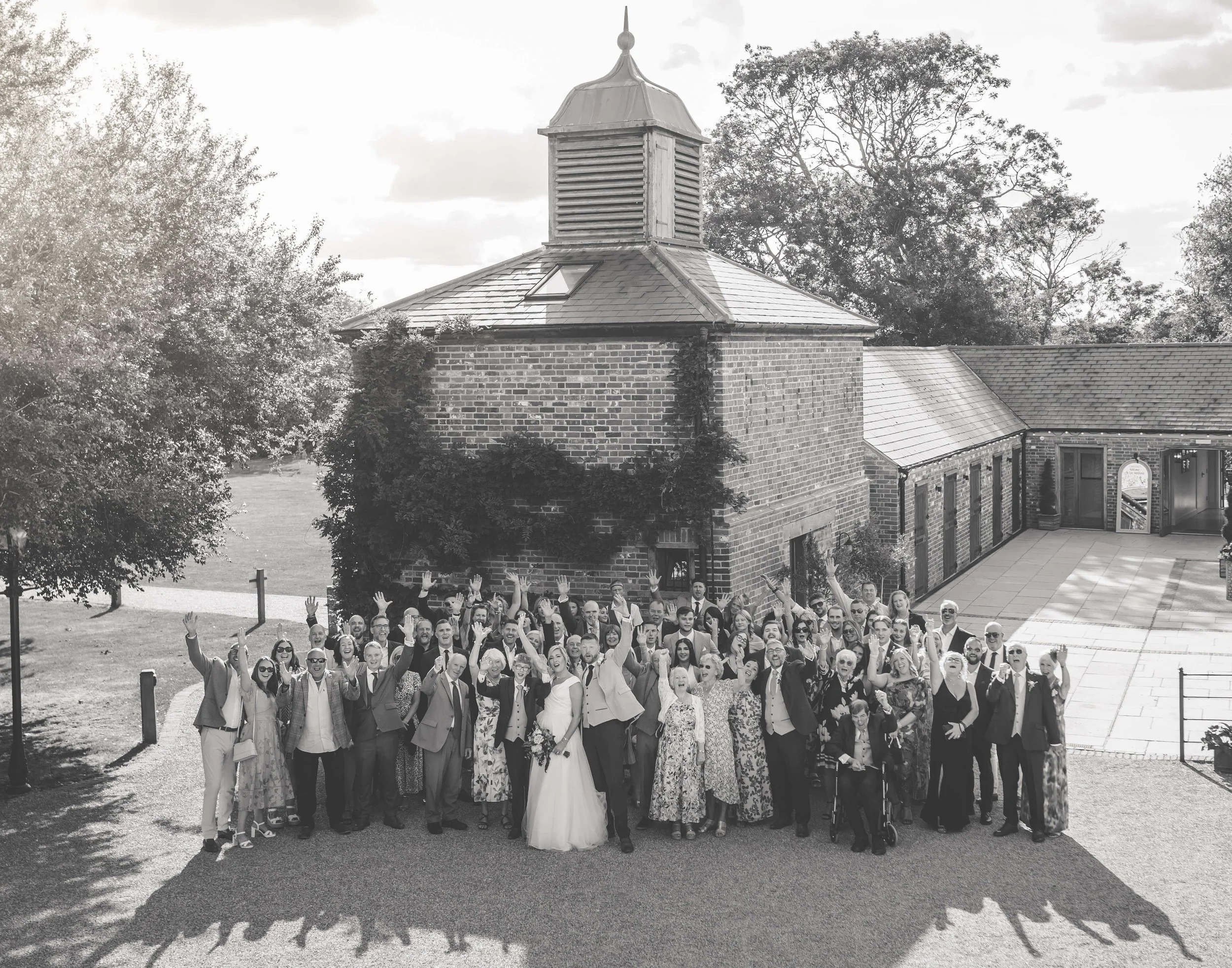 A large group of people dressed in formal attire gathered outside a brick building with a tower, celebrating a wedding.
