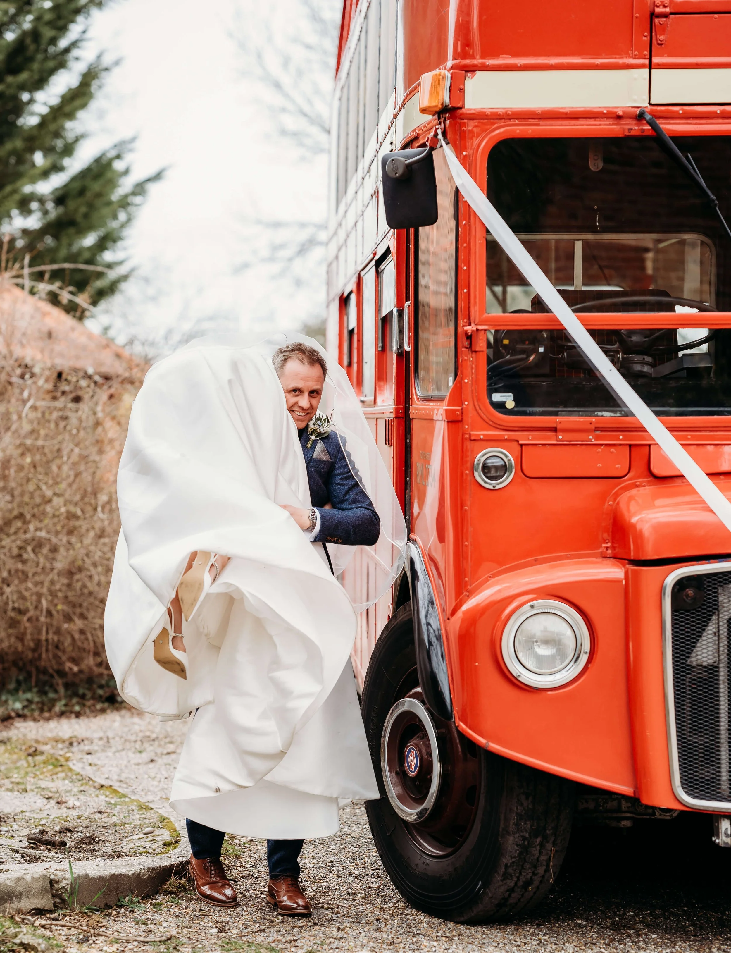 A man in formal attire is lifting his leg while standing next to a red double-decker bus, with the bus partially covering him.
