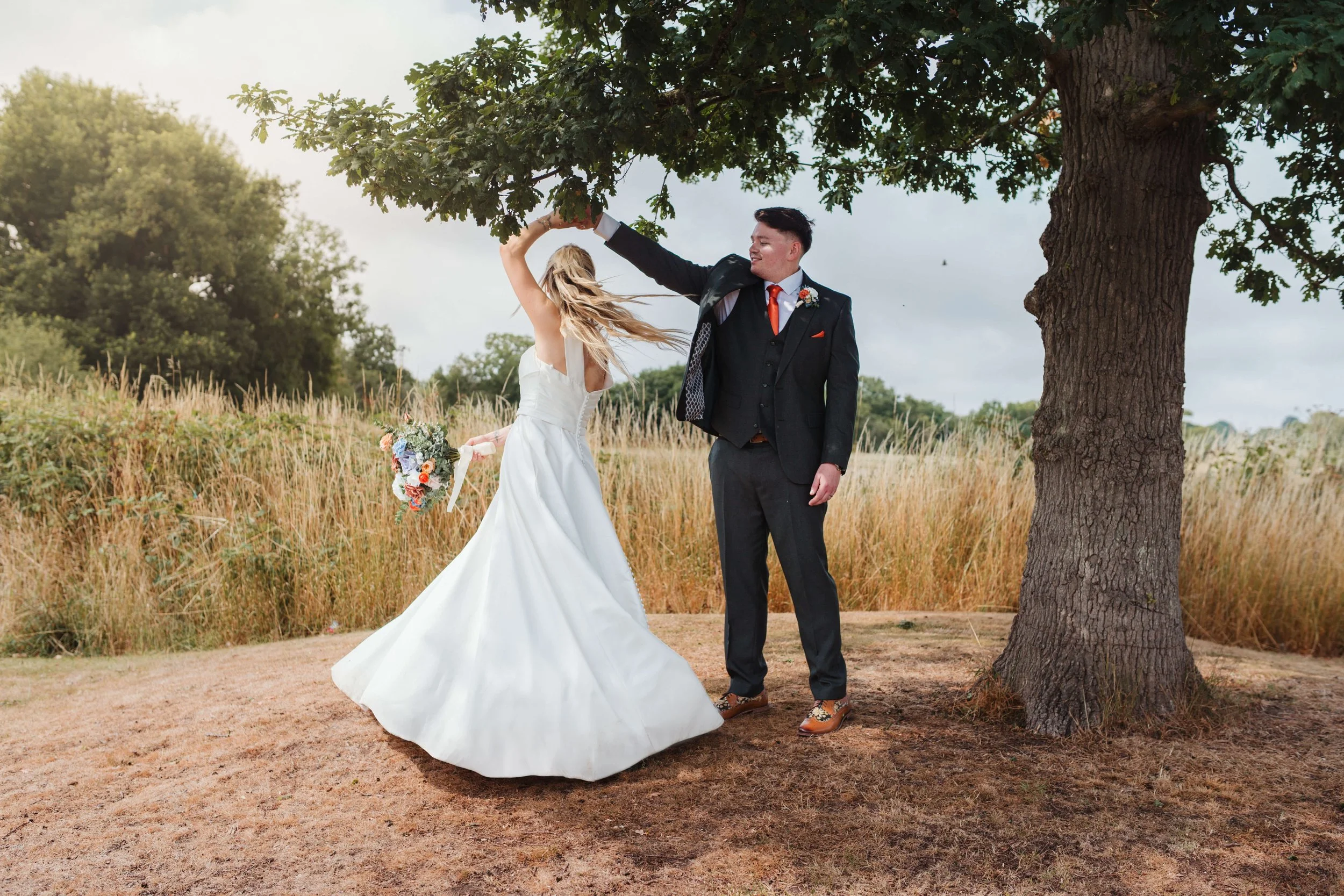 A bride in a white wedding dress holding a bouquet dancing with a groom in a black suit and red tie outdoors under a tree on a grassy field.