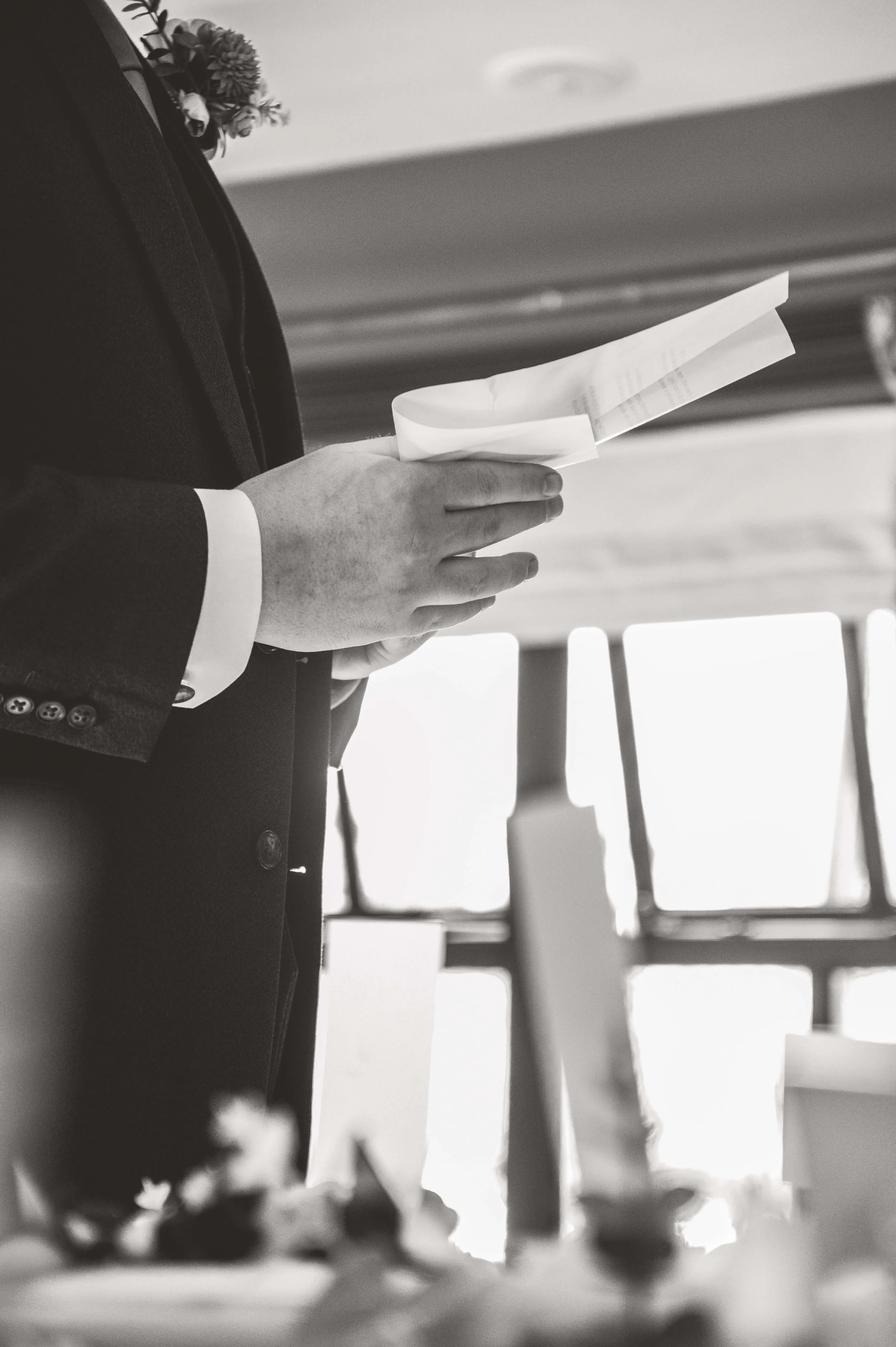 A person dressed in a suit holding and reading a piece of paper or program, with a blurred background that suggests an indoor setting with large windows.