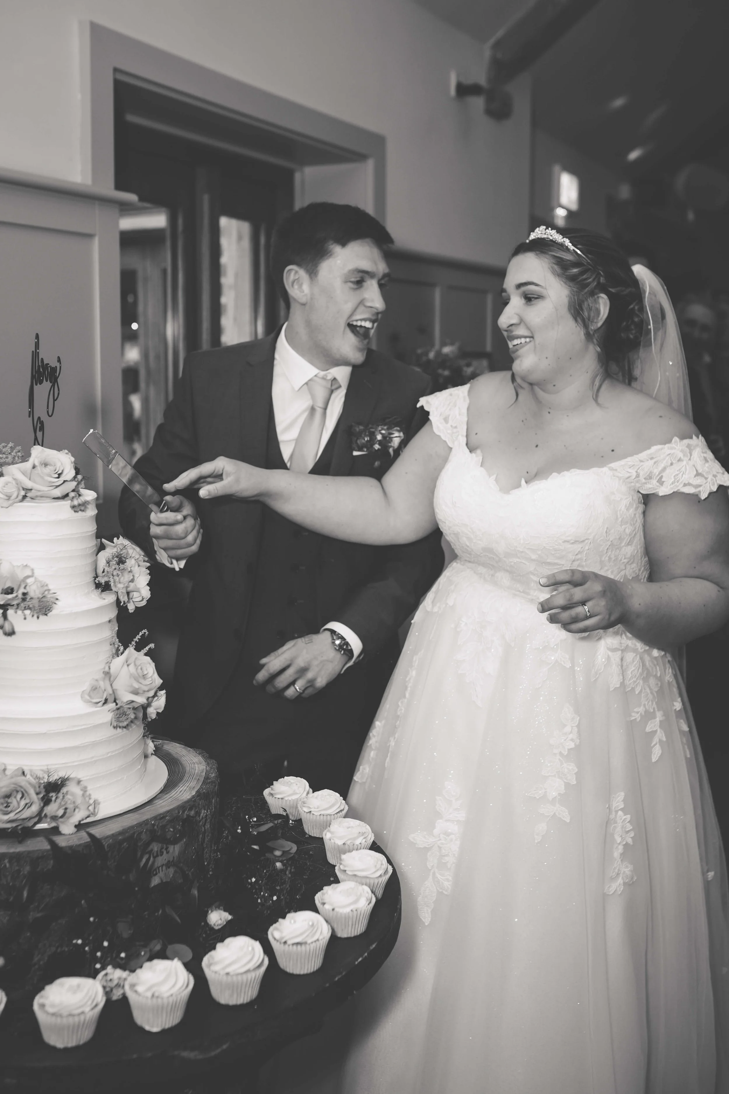 Black and white photo of a bride and groom at their wedding cake cutting, with the bride in a lace dress and a veil, and the groom in a suit, smiling.