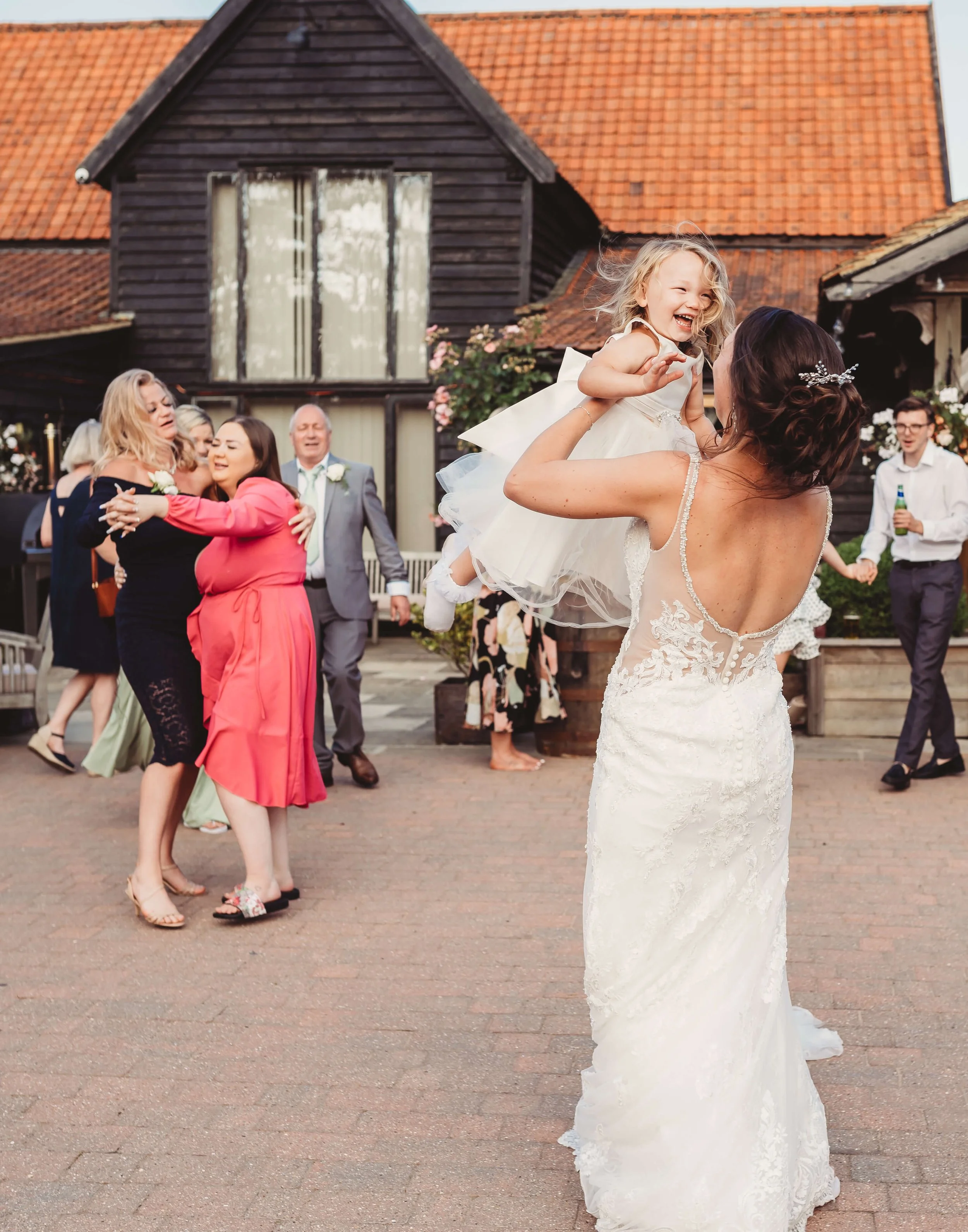 A bride holding a young girl in a white dress, smiling as they dance at a wedding reception outdoors with guests in the background.