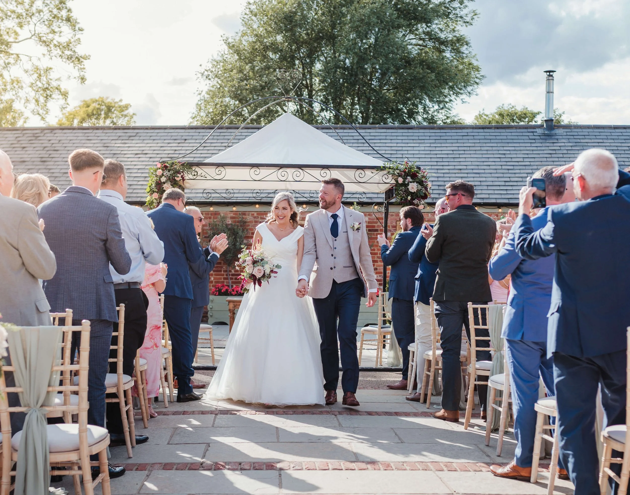 Couple walking down the aisle at an outdoor wedding ceremony, surrounded by guests clapping and taking photos, with a brick building and floral decorations in the background.