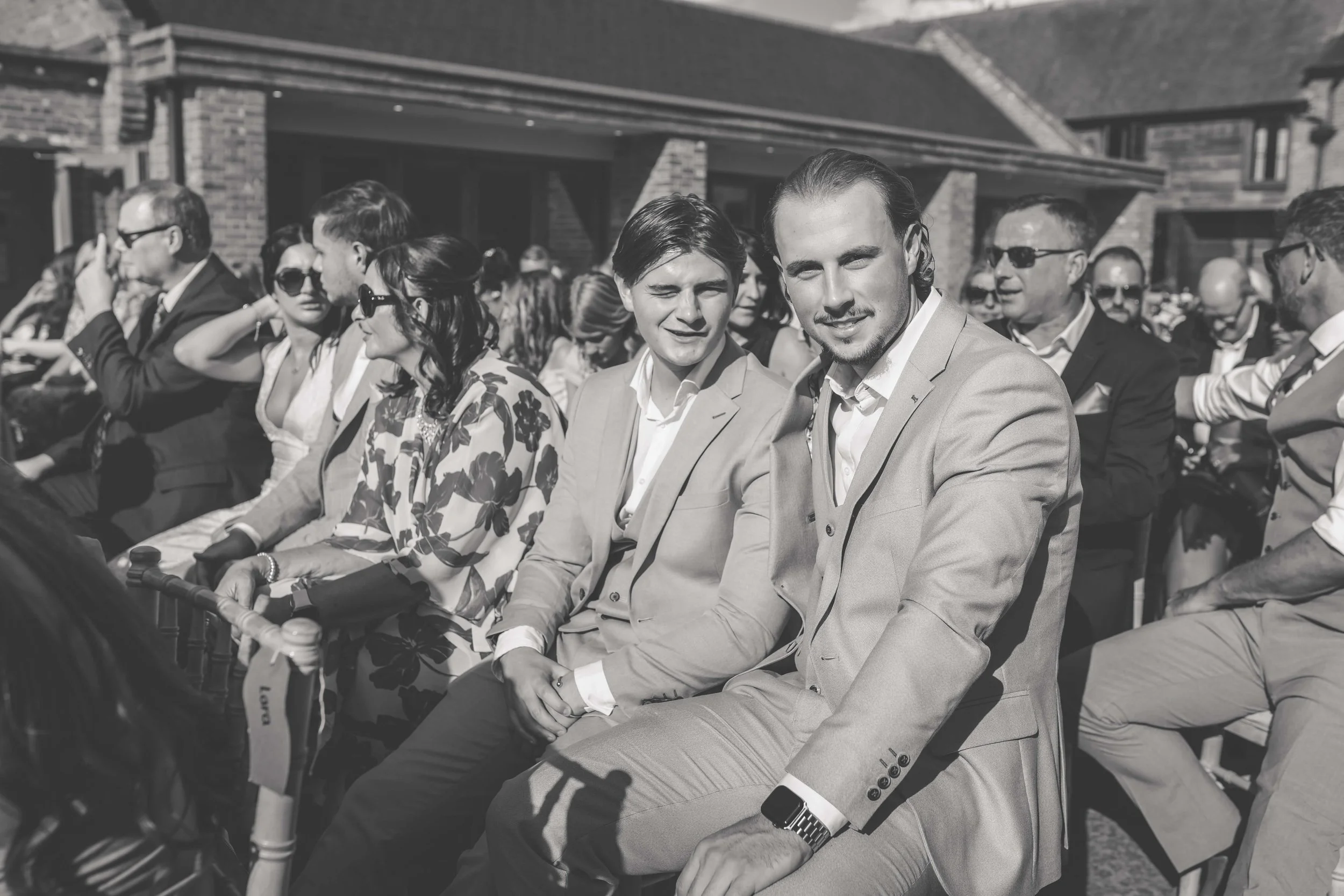 Black and white photo of people in formal attire sitting outdoors during daytime, possibly at a wedding or event, with a large brick building in the background.