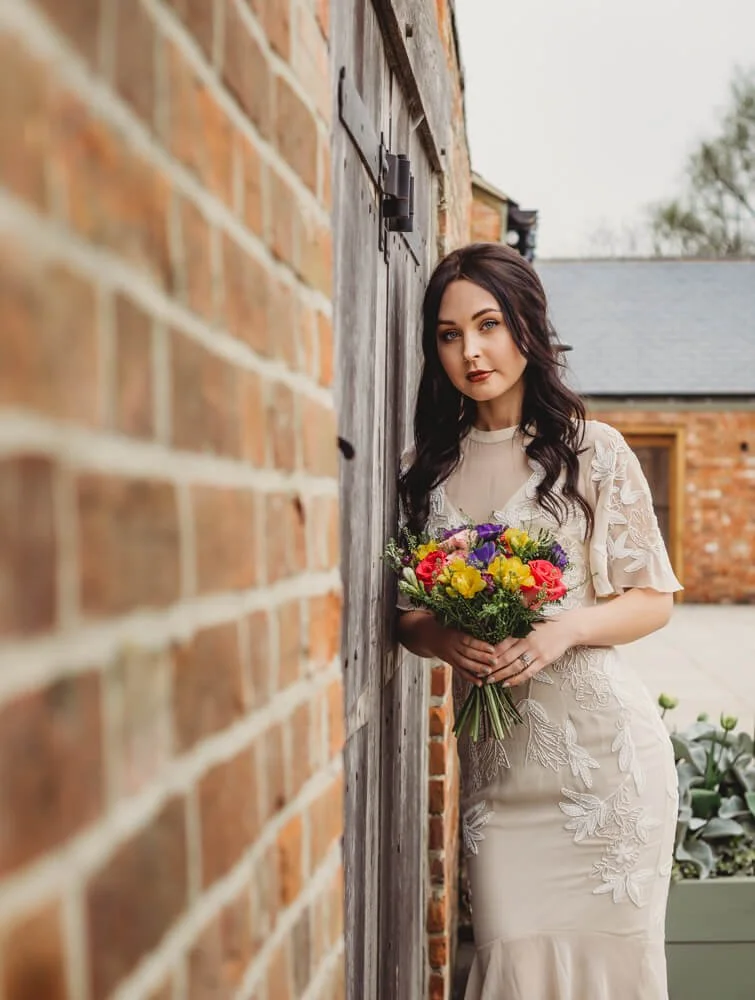 Woman in a cream dress holding a colorful bouquet of flowers, standing next to a brick wall outside.