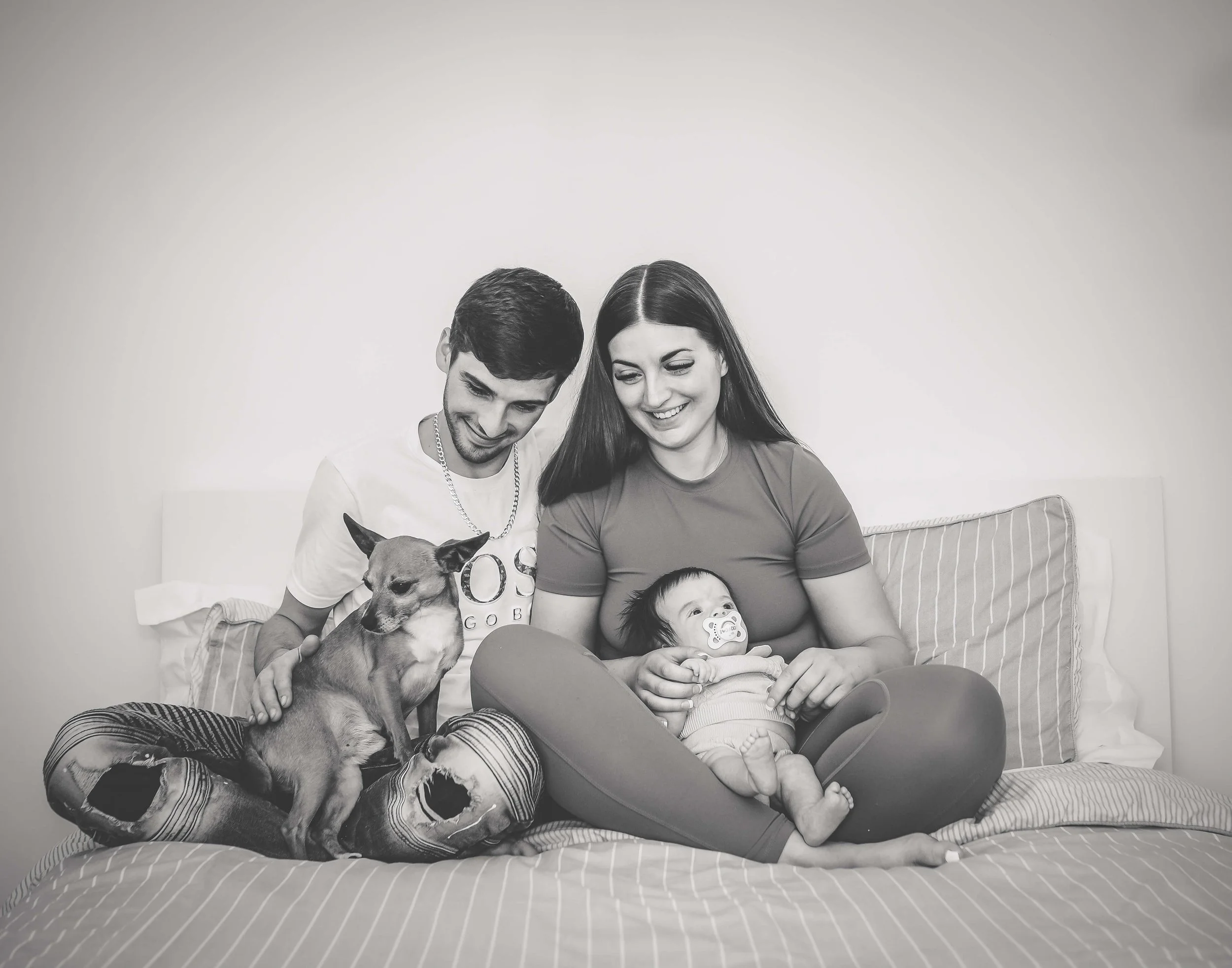 A happy family of four, including a man, woman, baby, and small dog, sitting on a bed together.