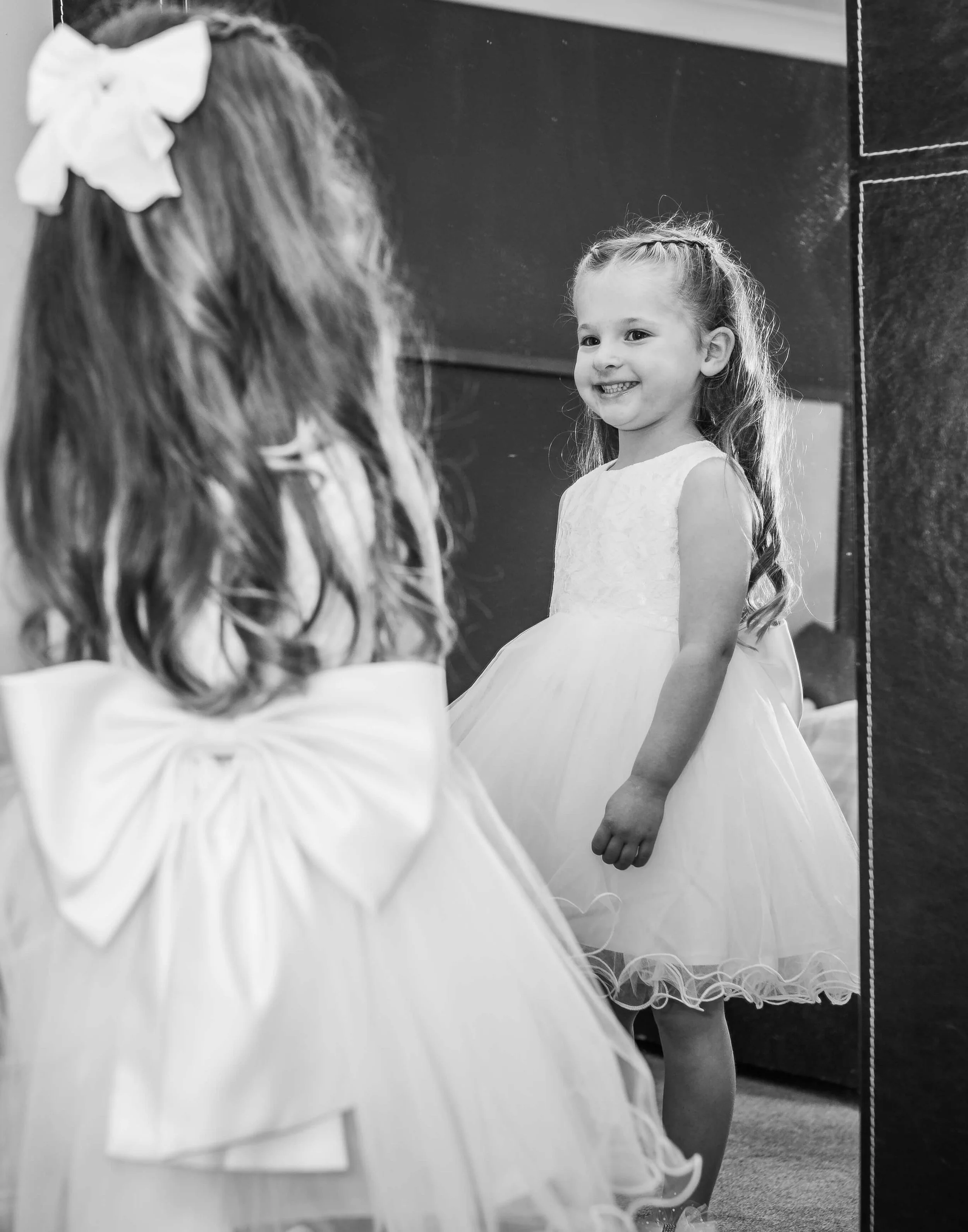 A young girl with long wavy hair, wearing a sleeveless dress, is looking at herself in a mirror and smiling. She is standing indoors.
