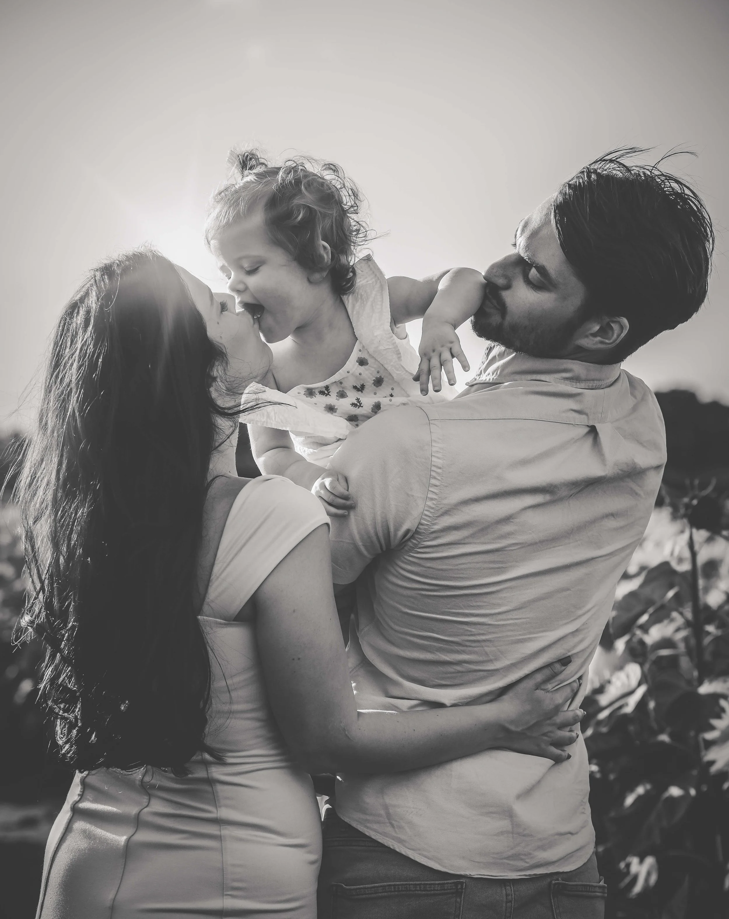 A happy family outdoors, with a man holding a young girl, and a woman kissing the girl, all smiling and enjoying a moment together.