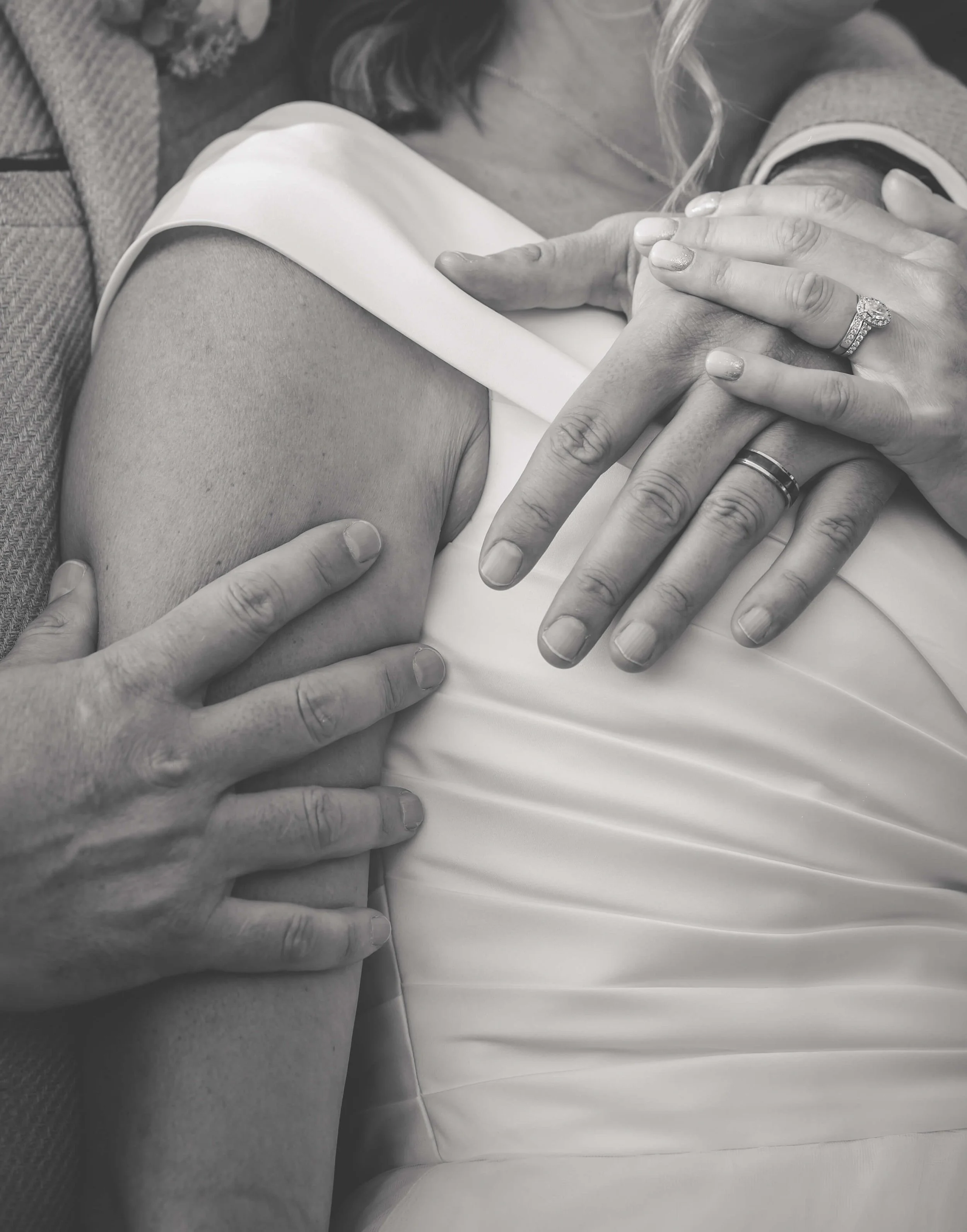 Close-up of hands gently holding a woman's shoulder and chest area, with rings on some fingers, in black-and-white photography.